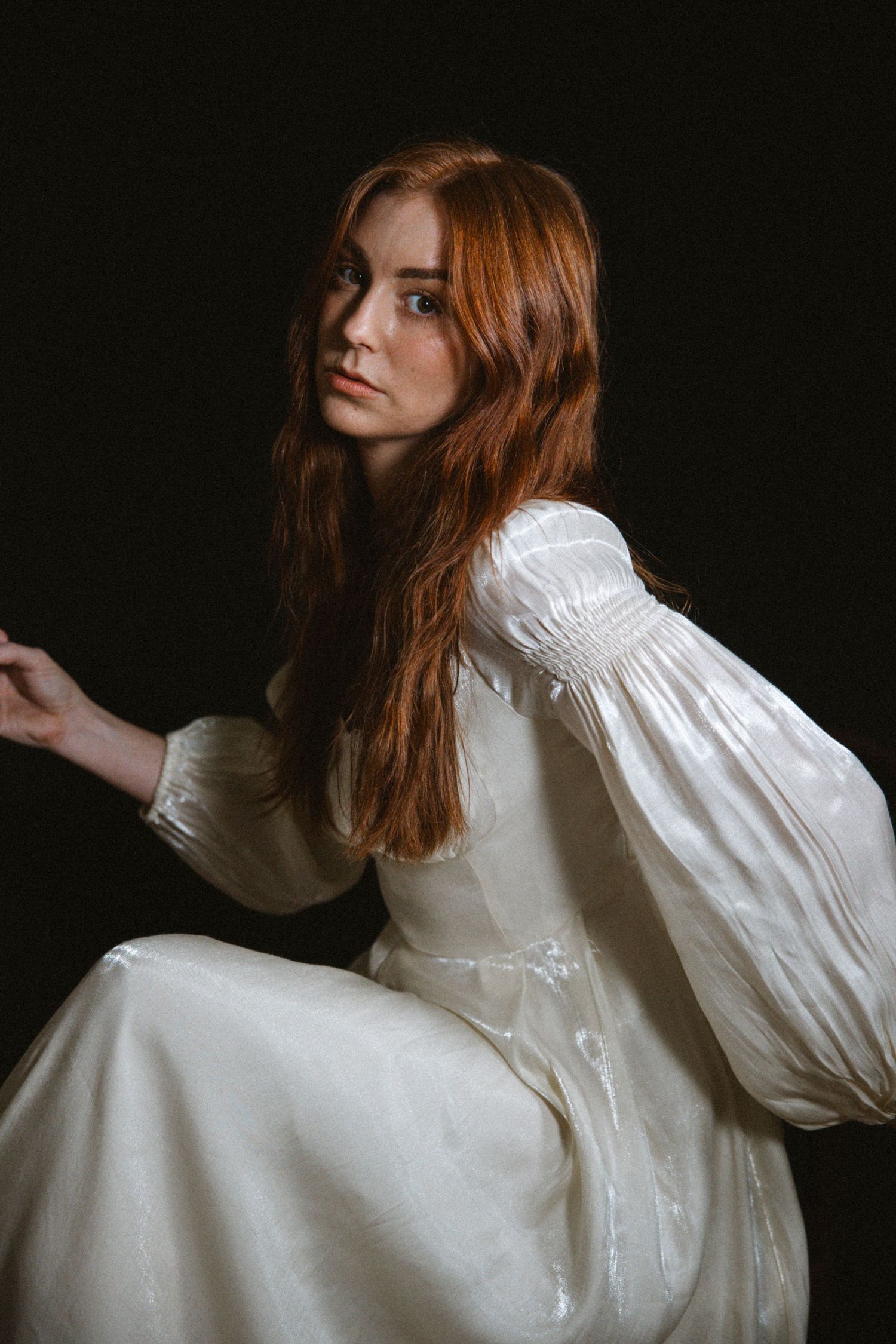 Artistic portrait in white vintage dress against dark background with dramatic lighting and flowing red hair.