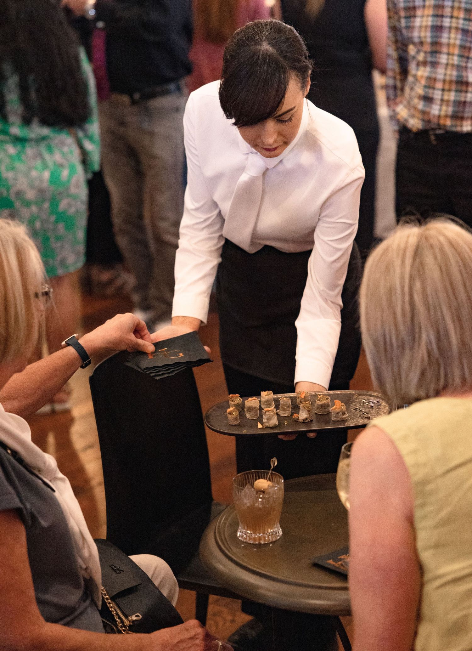 Server in white shirt presents appetizers on a tray to guests at an elegant dining event.