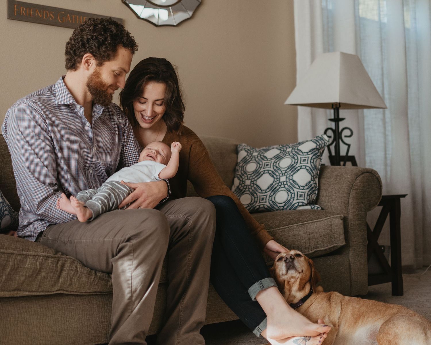 Couple with newborn baby relaxing on brown couch with dog in cozy living room.