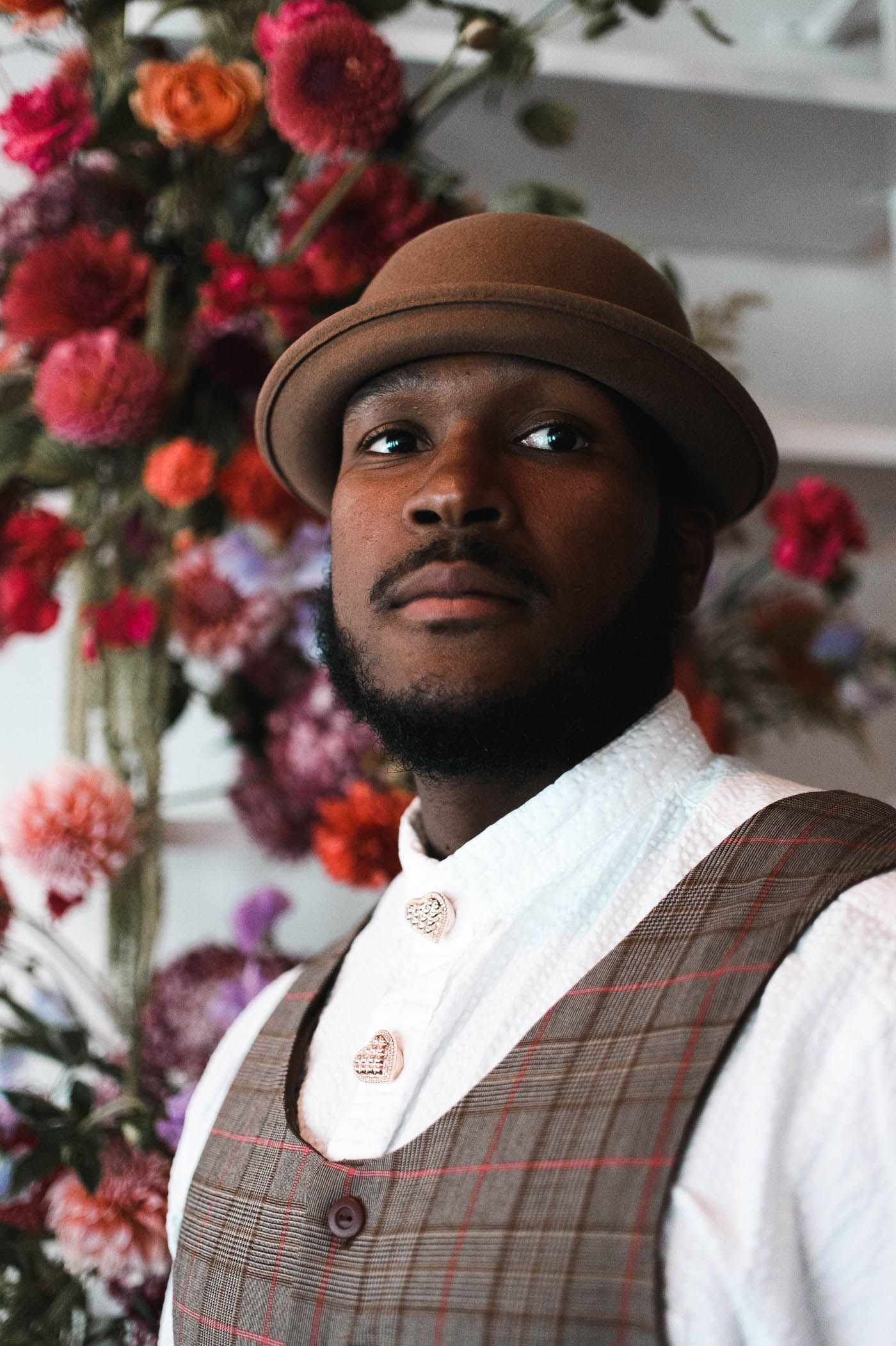 Portrait of a stylish person in a brown hat and vest against a floral backdrop.