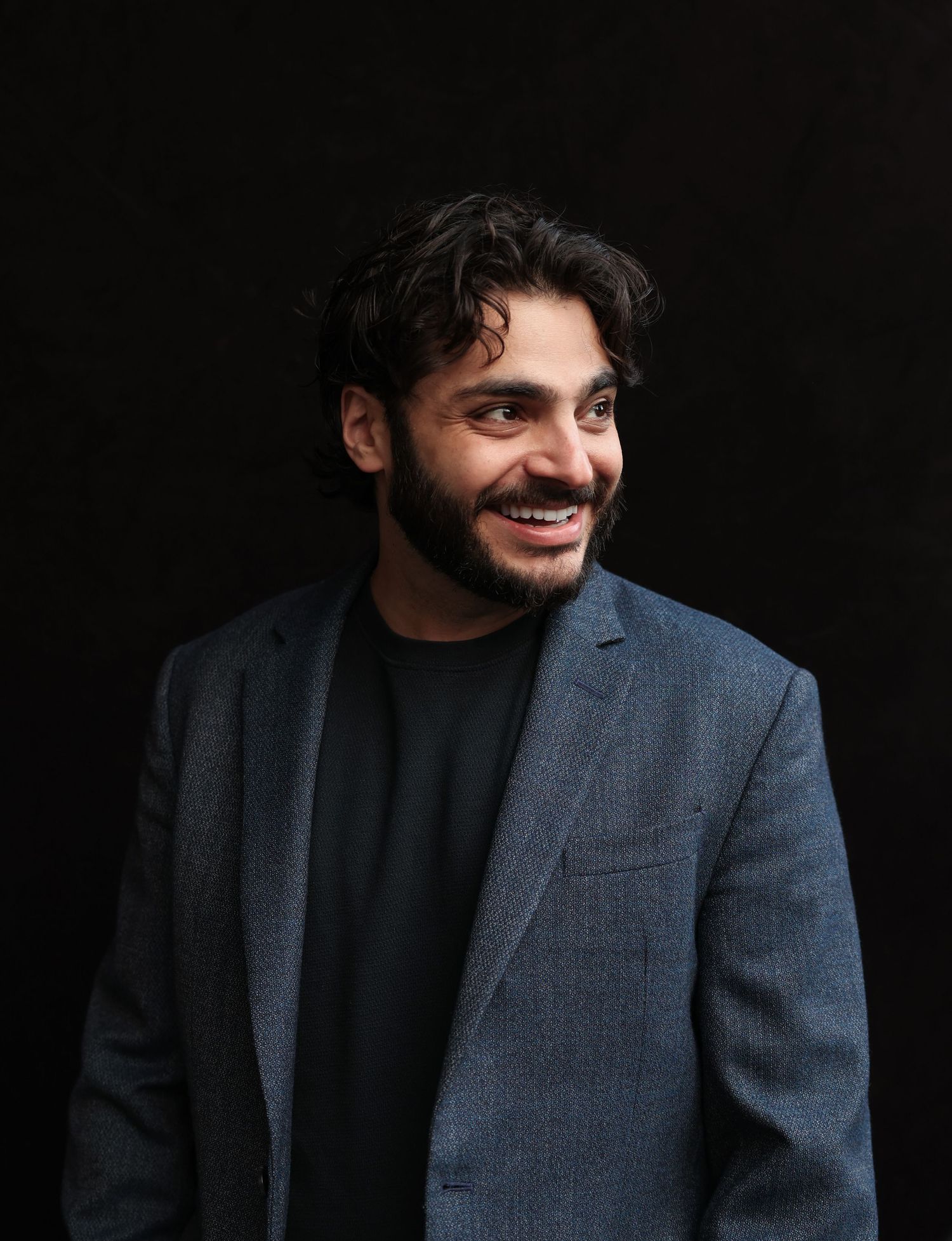 Professional headshot of person in dark blazer and black shirt against black background smiling warmly.