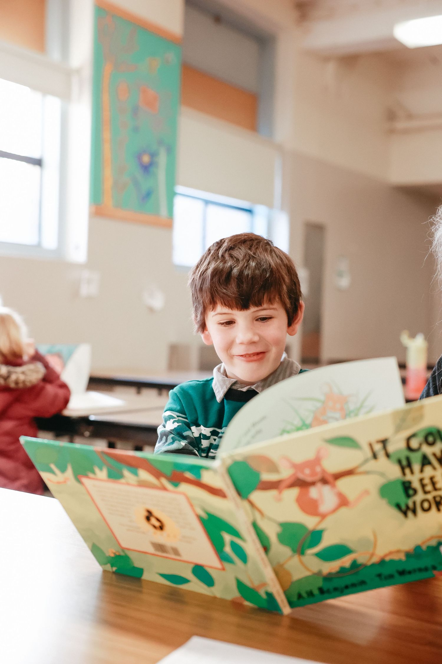 A student smiles while looking at a colorful pop-up book with green and orange illustrations on a wooden table.