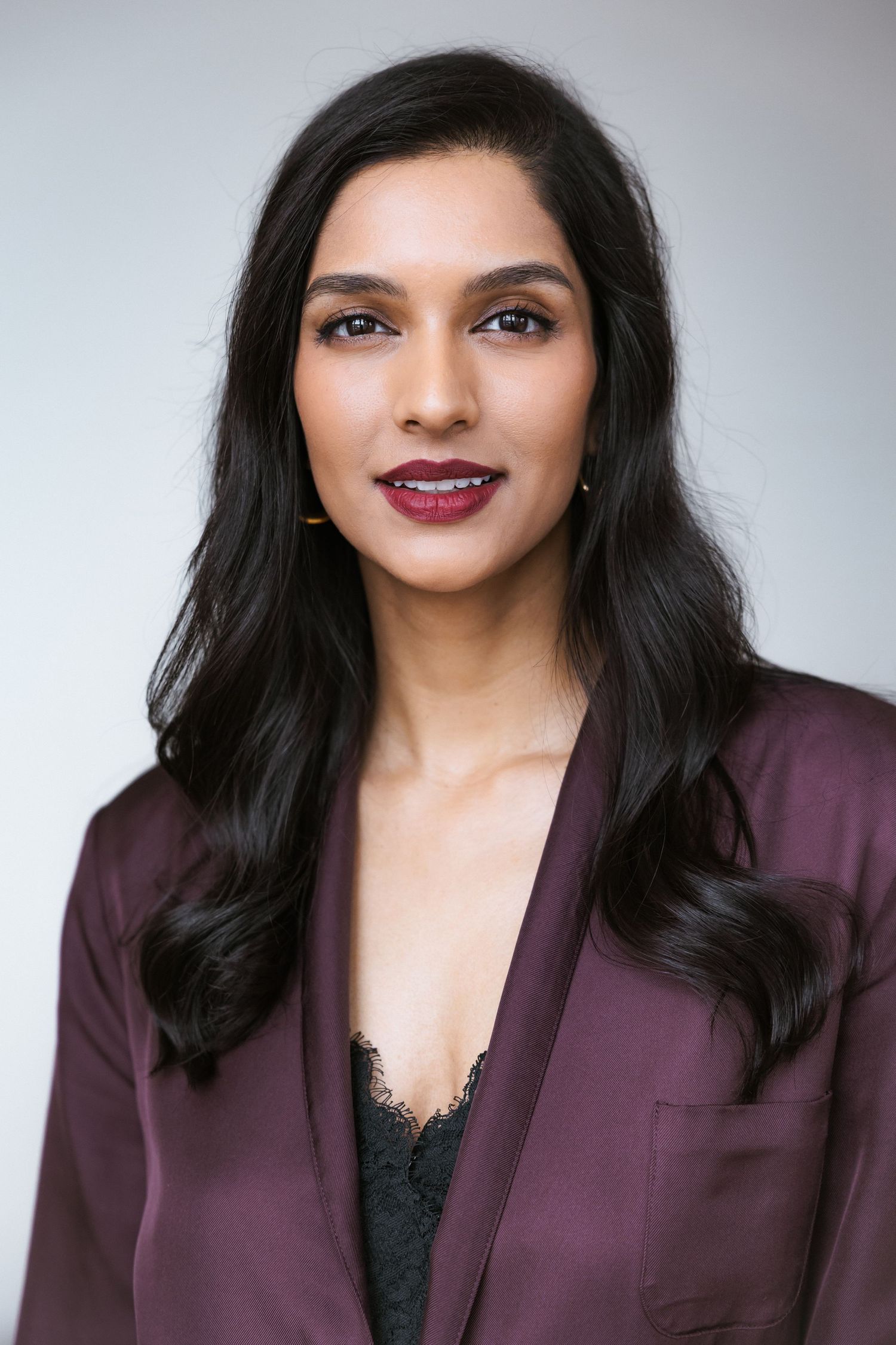 Professional headshot of a young South Asian woman in a burgundy blazer with dark wavy hair and bold red lipstick.
