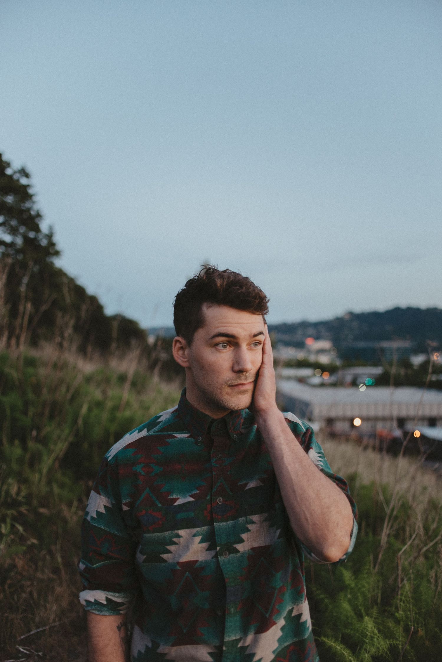 A person wearing a patterned shirt sits thoughtfully in a coastal outdoor setting at dusk.