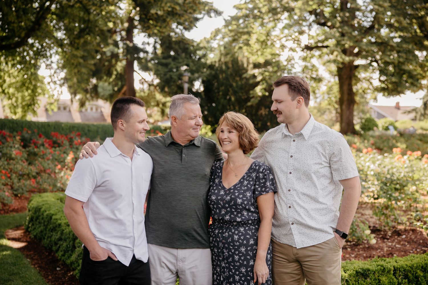 A family group poses together outdoors in front of blooming rose bushes and trees on a sunny day.