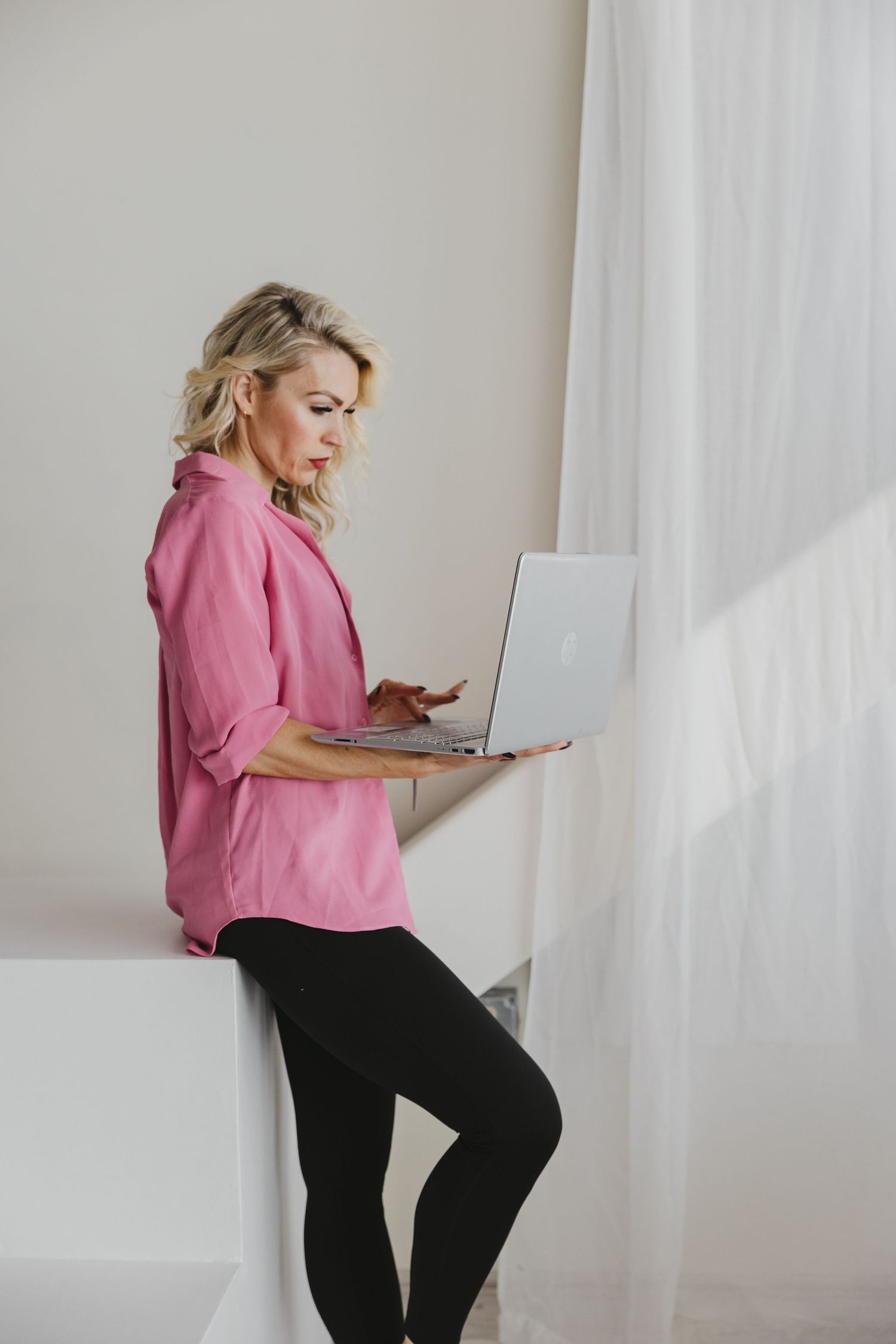 Person in pink shirt and black pants using laptop while sitting casually against white wall in minimalist setting.