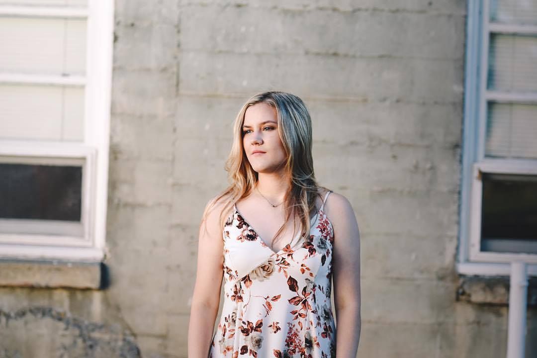 Woman in floral dress poses against industrial concrete wall background.