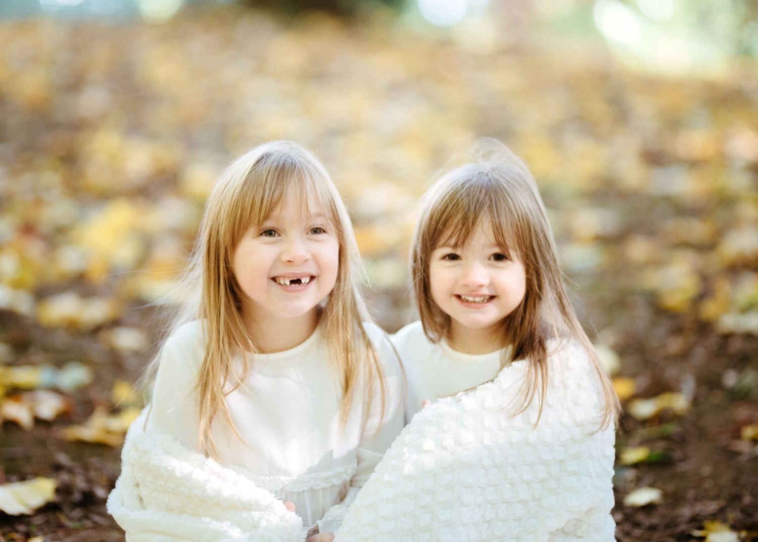 Two young children wrapped in white blankets smiling among autumn leaves.