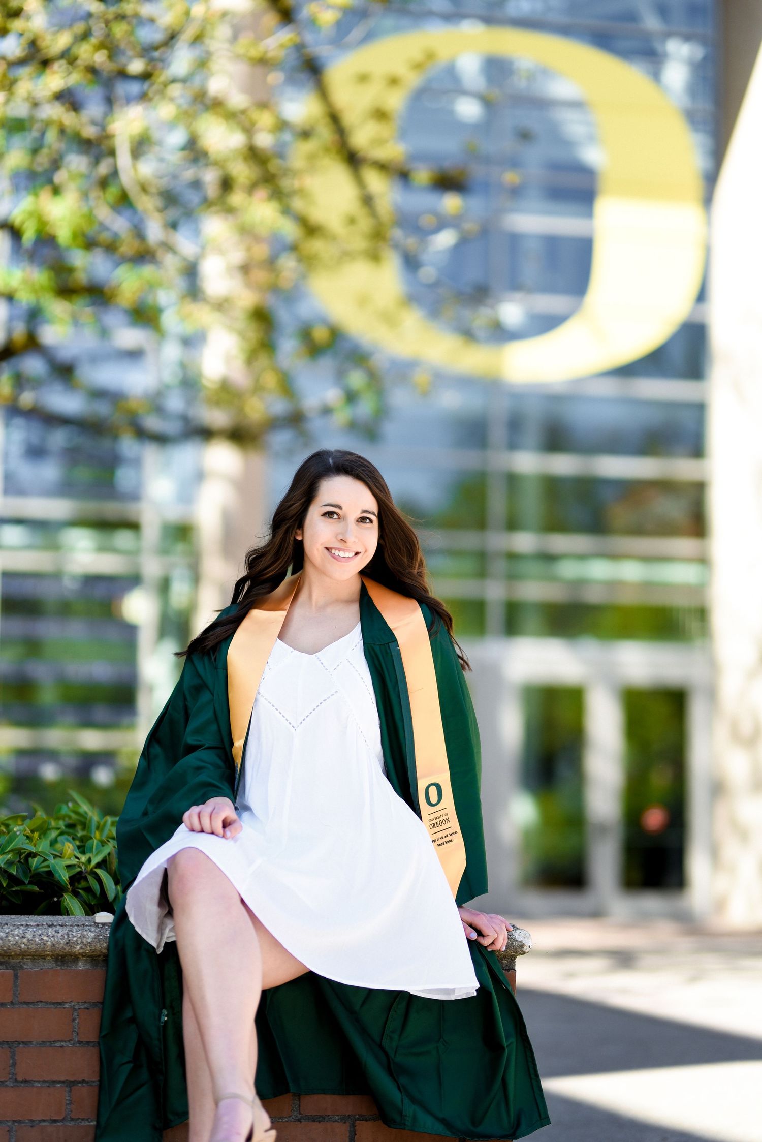 Graduate in white dress and green gown near Oregon campus logo.