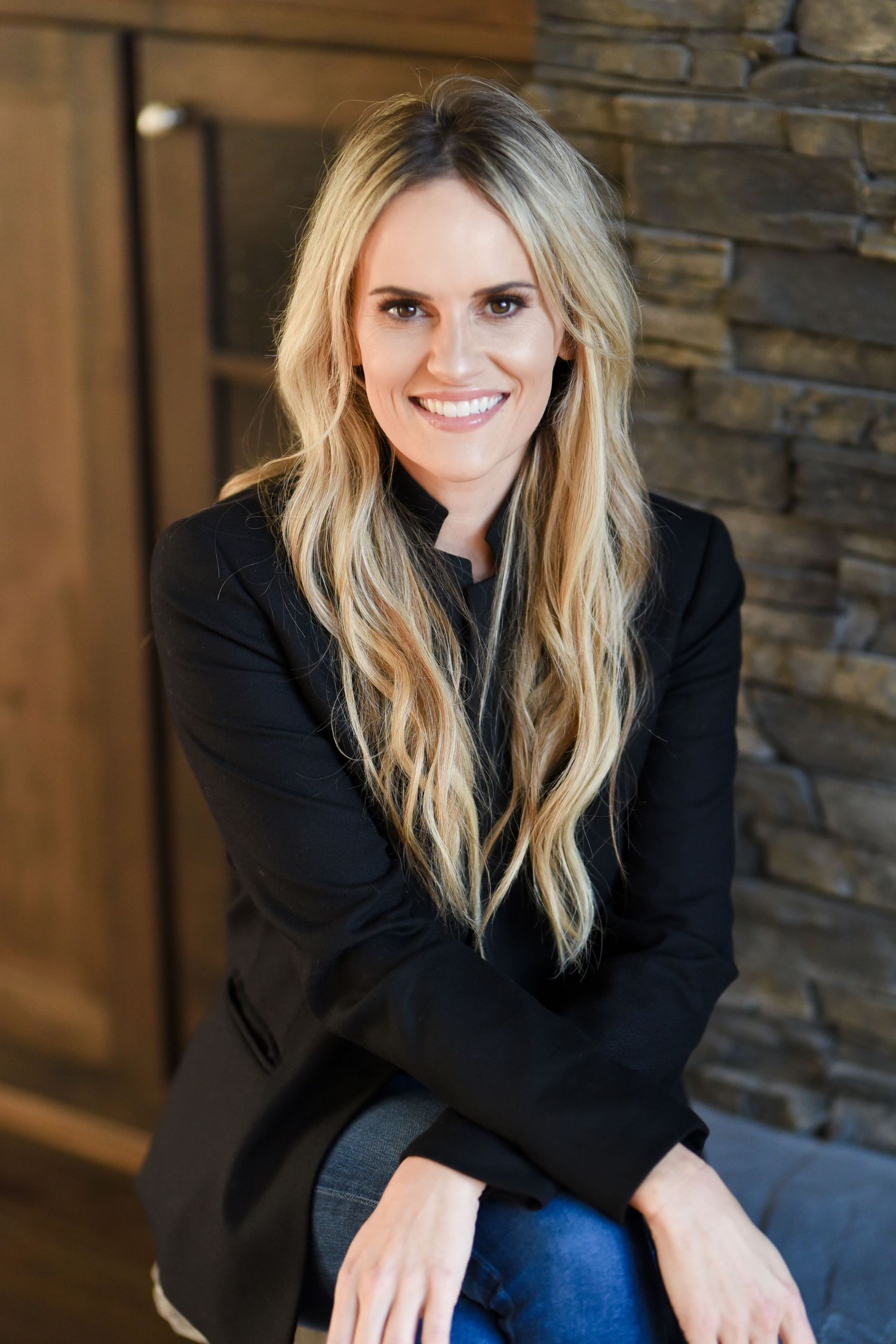 Professional headshot of a person wearing a black blazer and jeans in front of a stone wall.