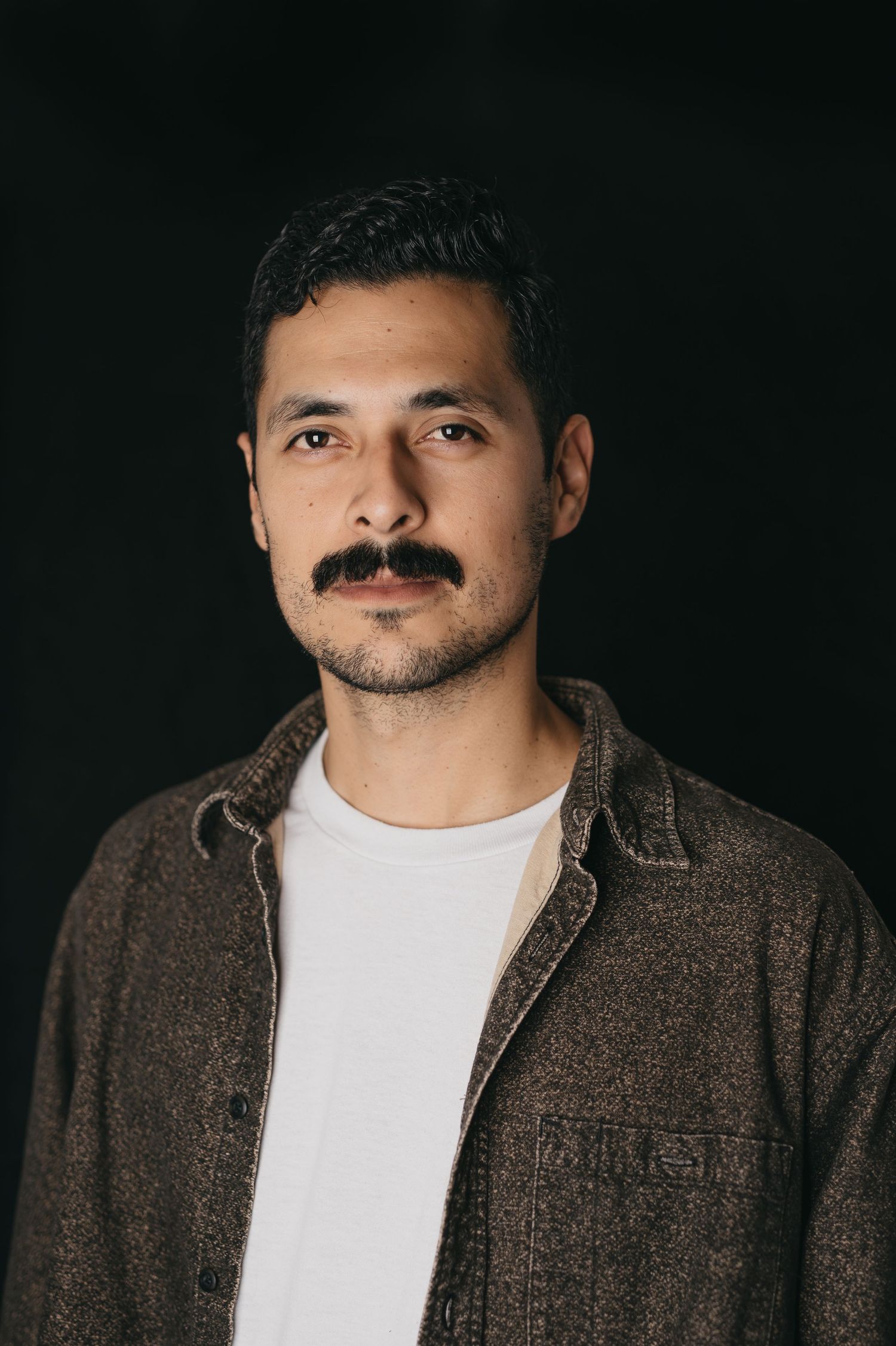 Studio portrait of person wearing brown jacket and white t-shirt against dark background with dramatic lighting.