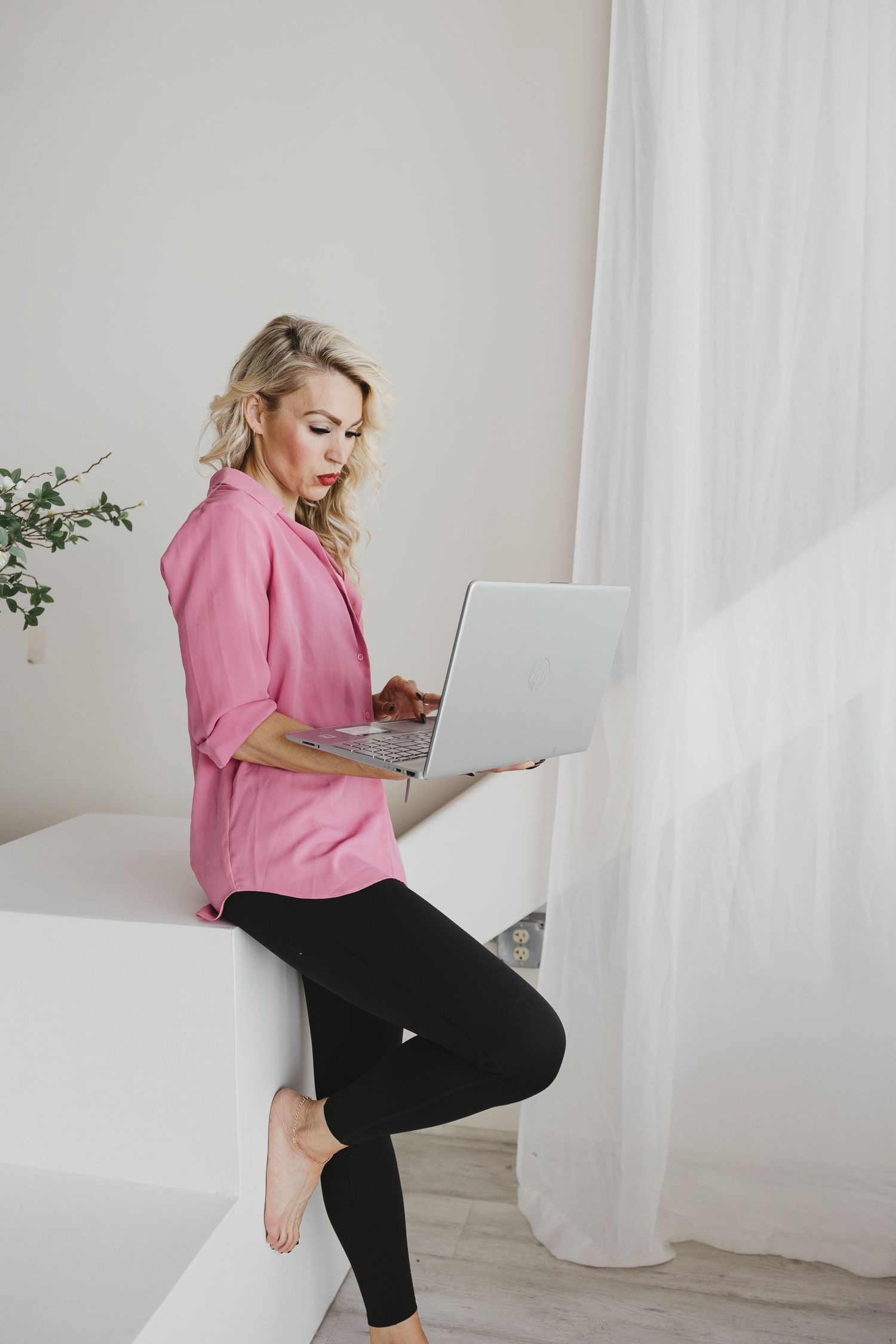 Woman in pink blazer and black leggings working on laptop while sitting casually on white surface near curtain.