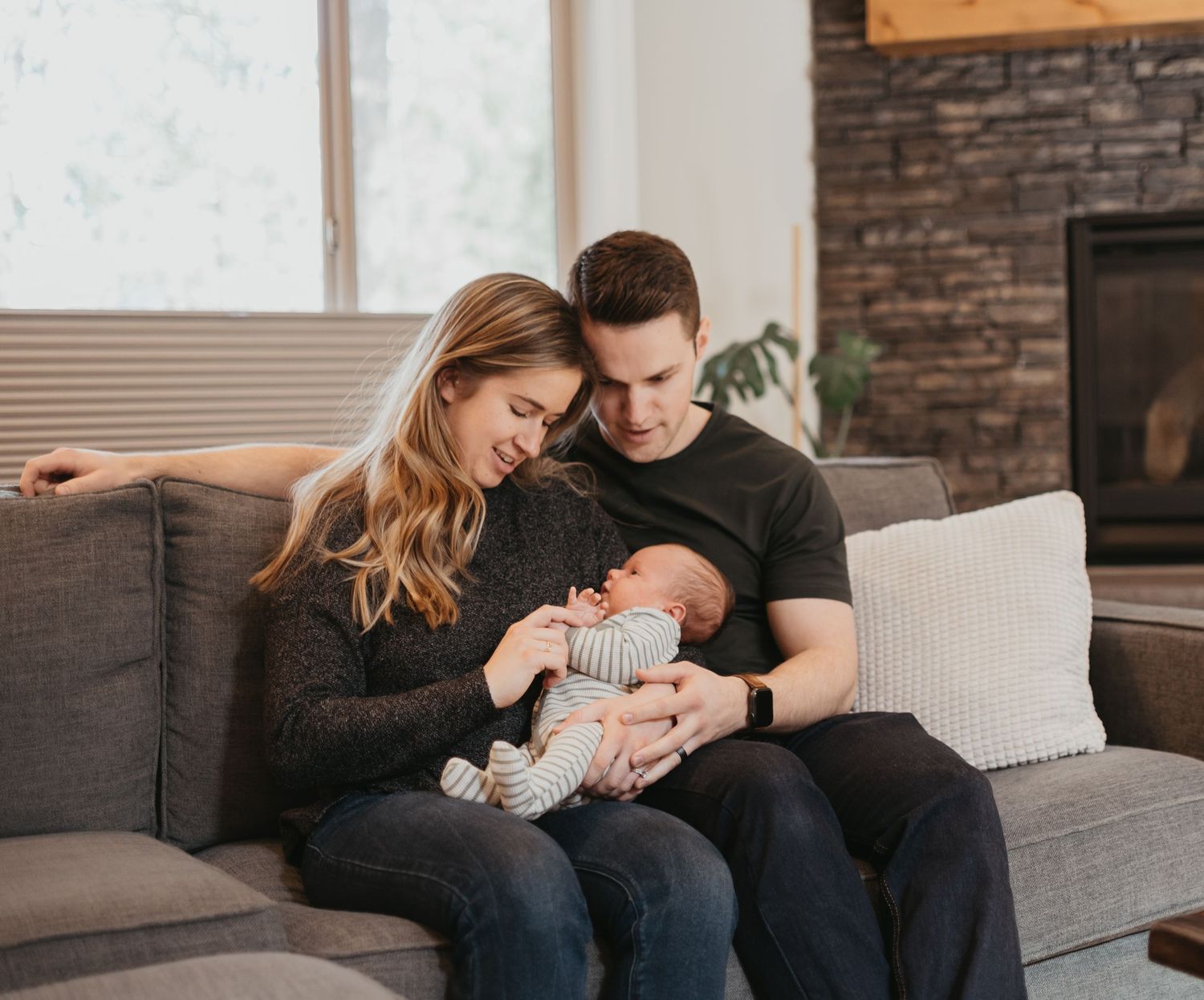 Parents cuddle with newborn baby on gray couch in modern living room.