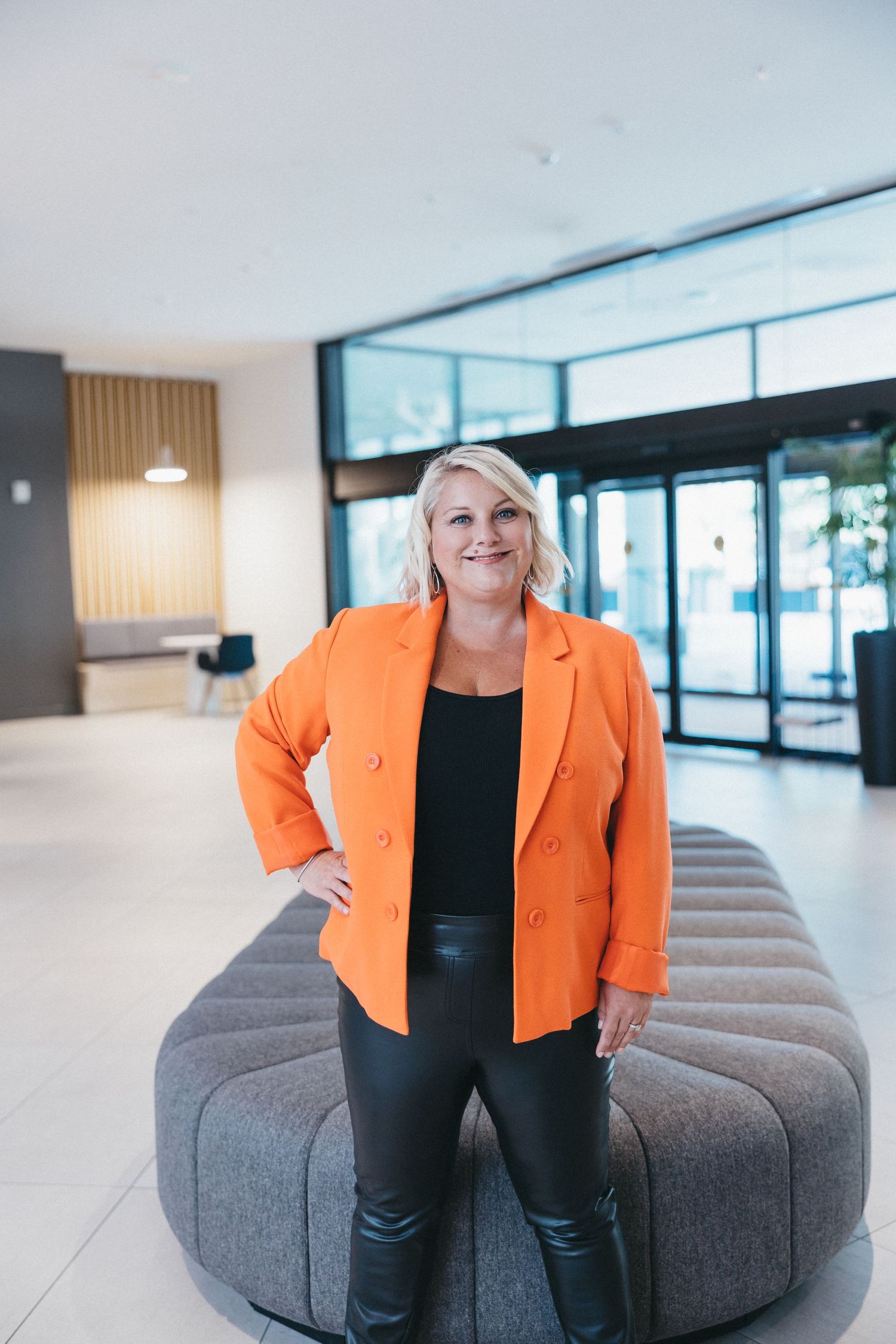 Professional in bright orange blazer and black outfit seated on modern gray couch in bright office setting.
