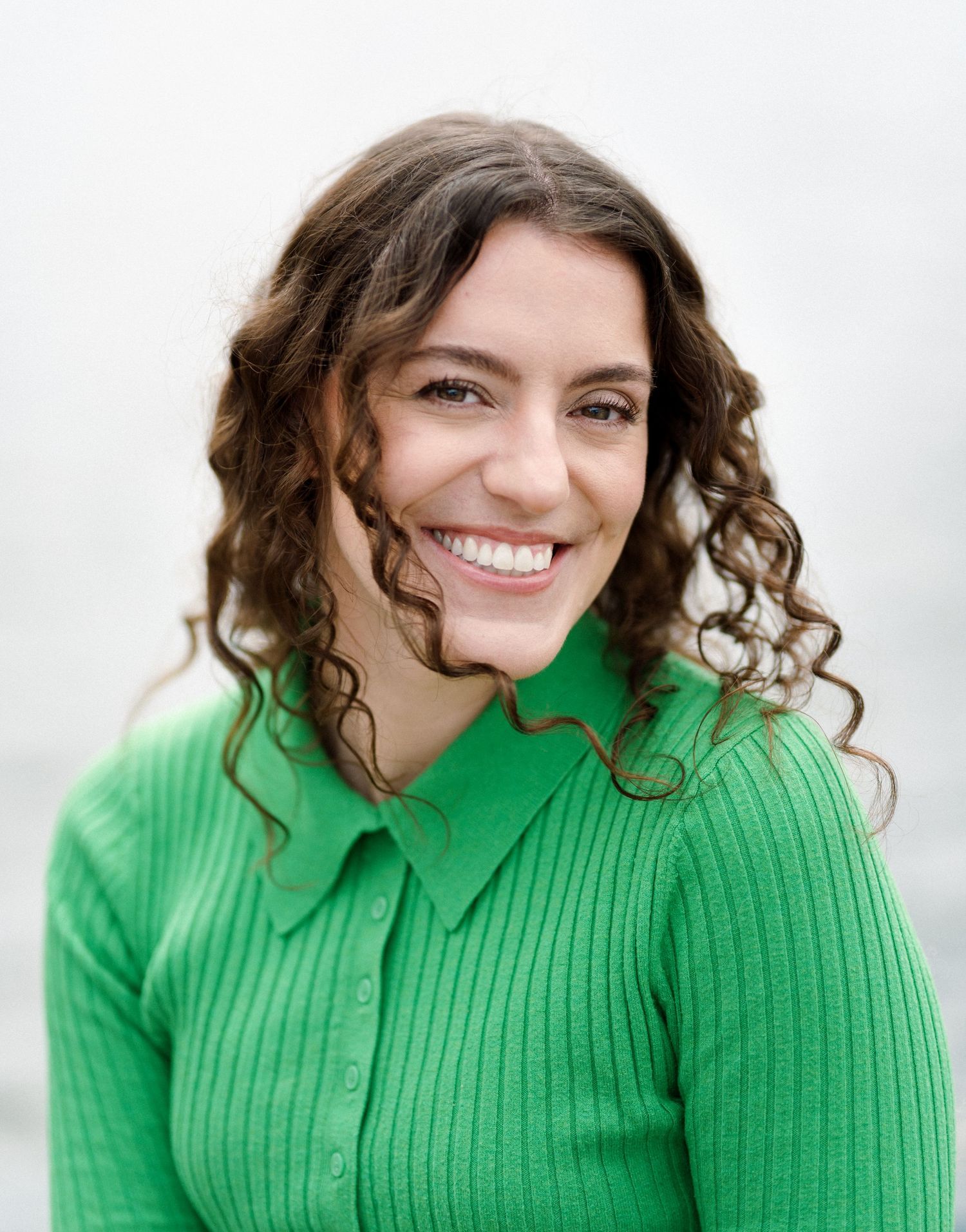 Smiling woman with curly brown hair wearing a green ribbed collared sweater against a light background.