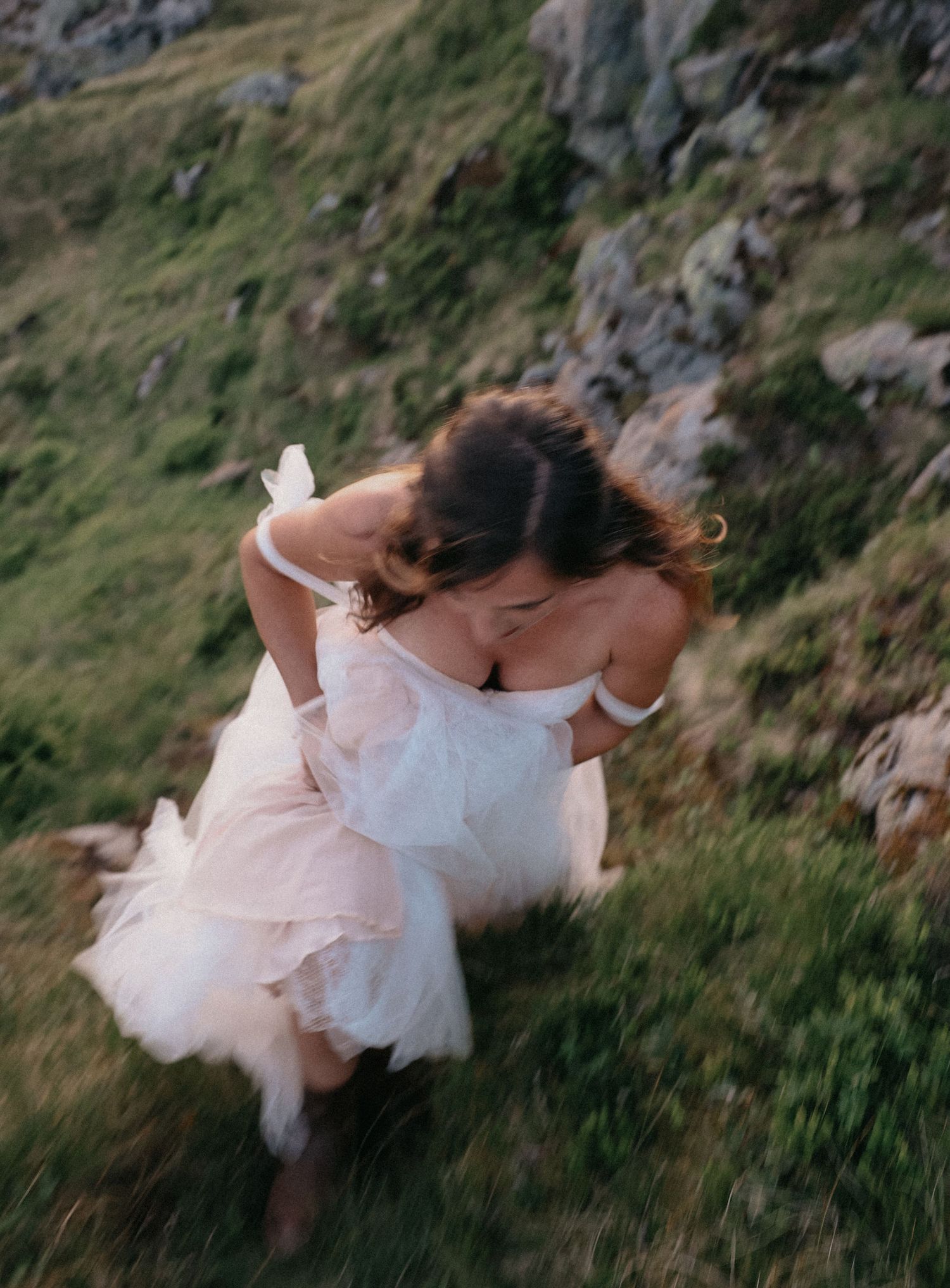 A bride in a flowing white dress twirls on a mountainside path surrounded by natural scenery.