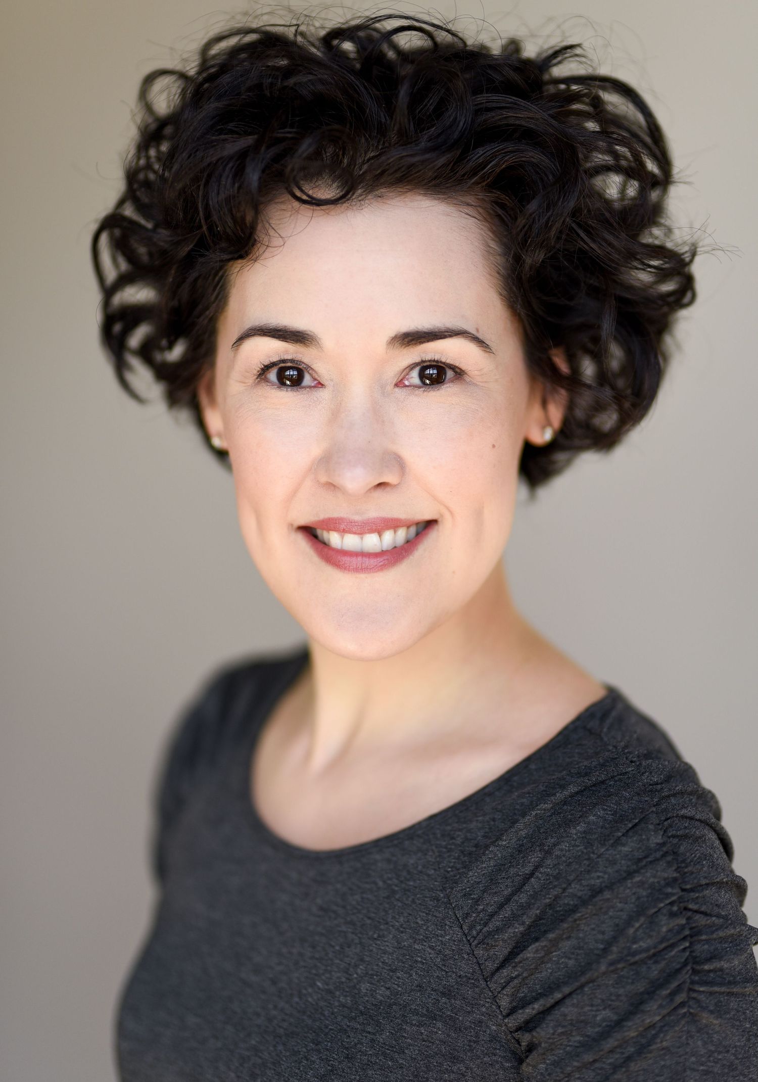 Studio portrait featuring dark curly hair and natural makeup against a neutral background.