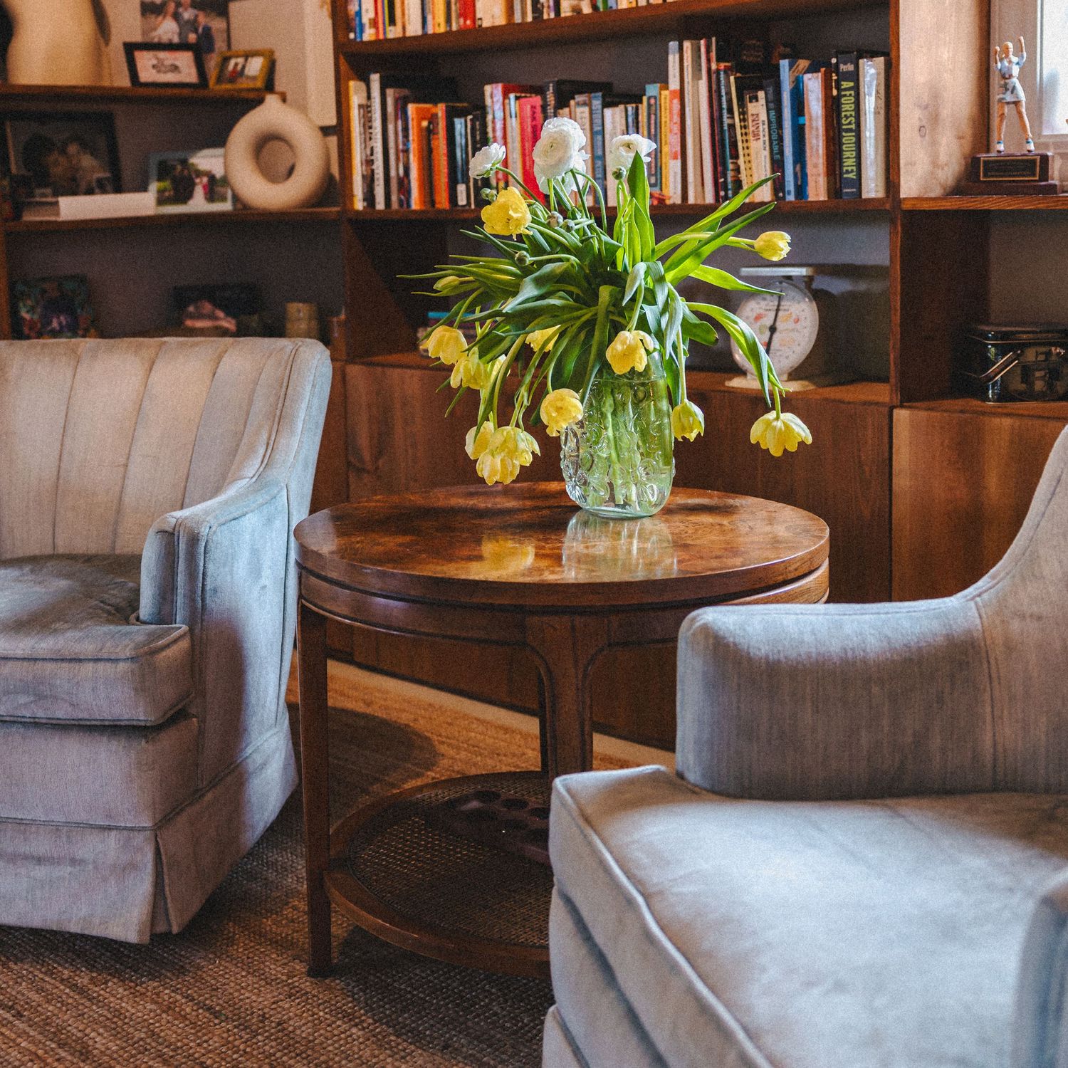 Elegant sitting area with gray armchairs and yellow tulips in glass vase on round wooden table.