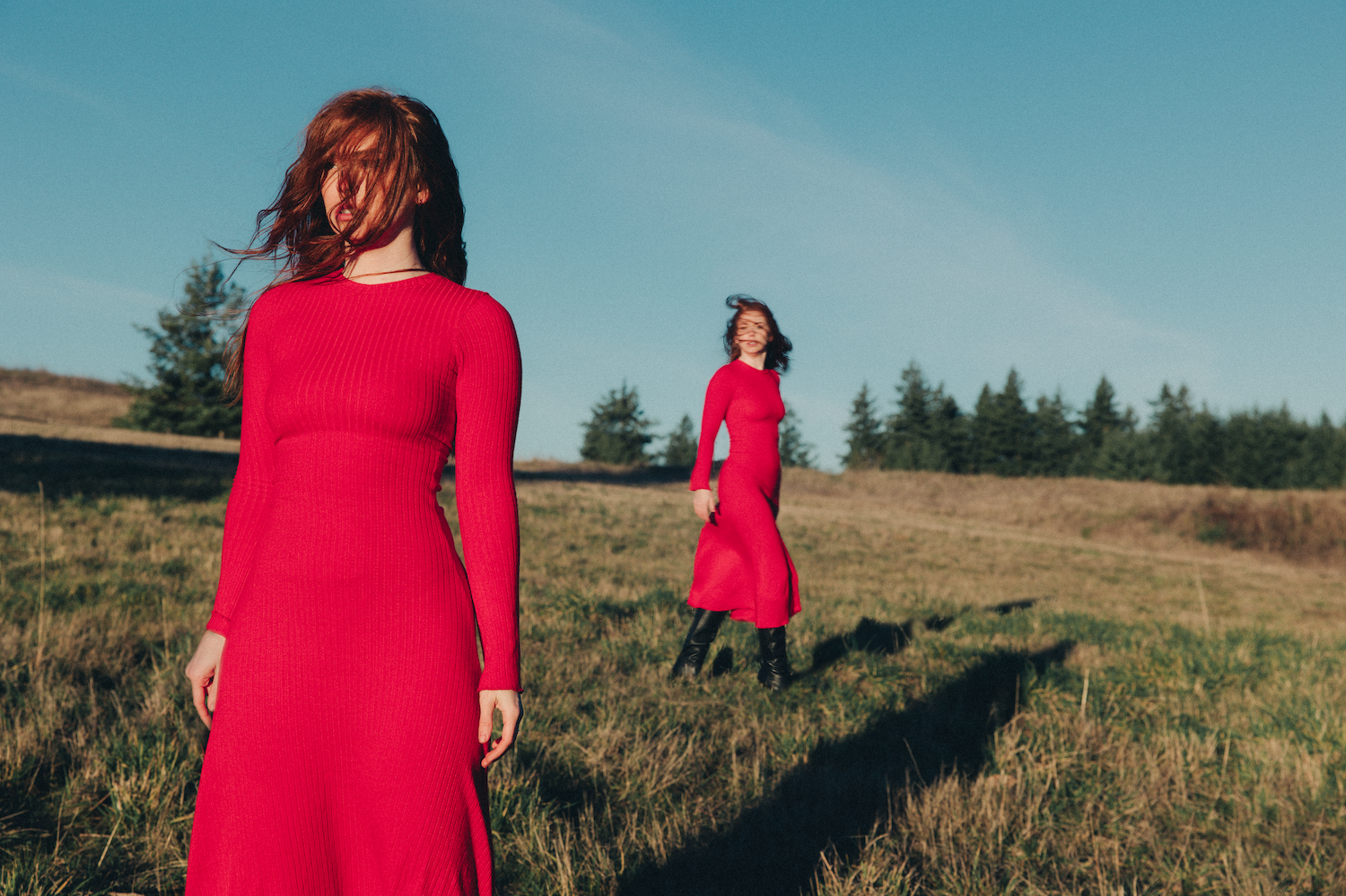 Two women in bright red dresses stand in a golden field with evergreen trees in the background on a sunny day.