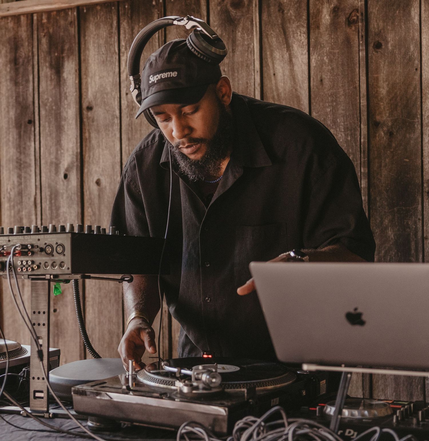 DJ performs with turntables and laptop setup against rustic wooden wall backdrop.