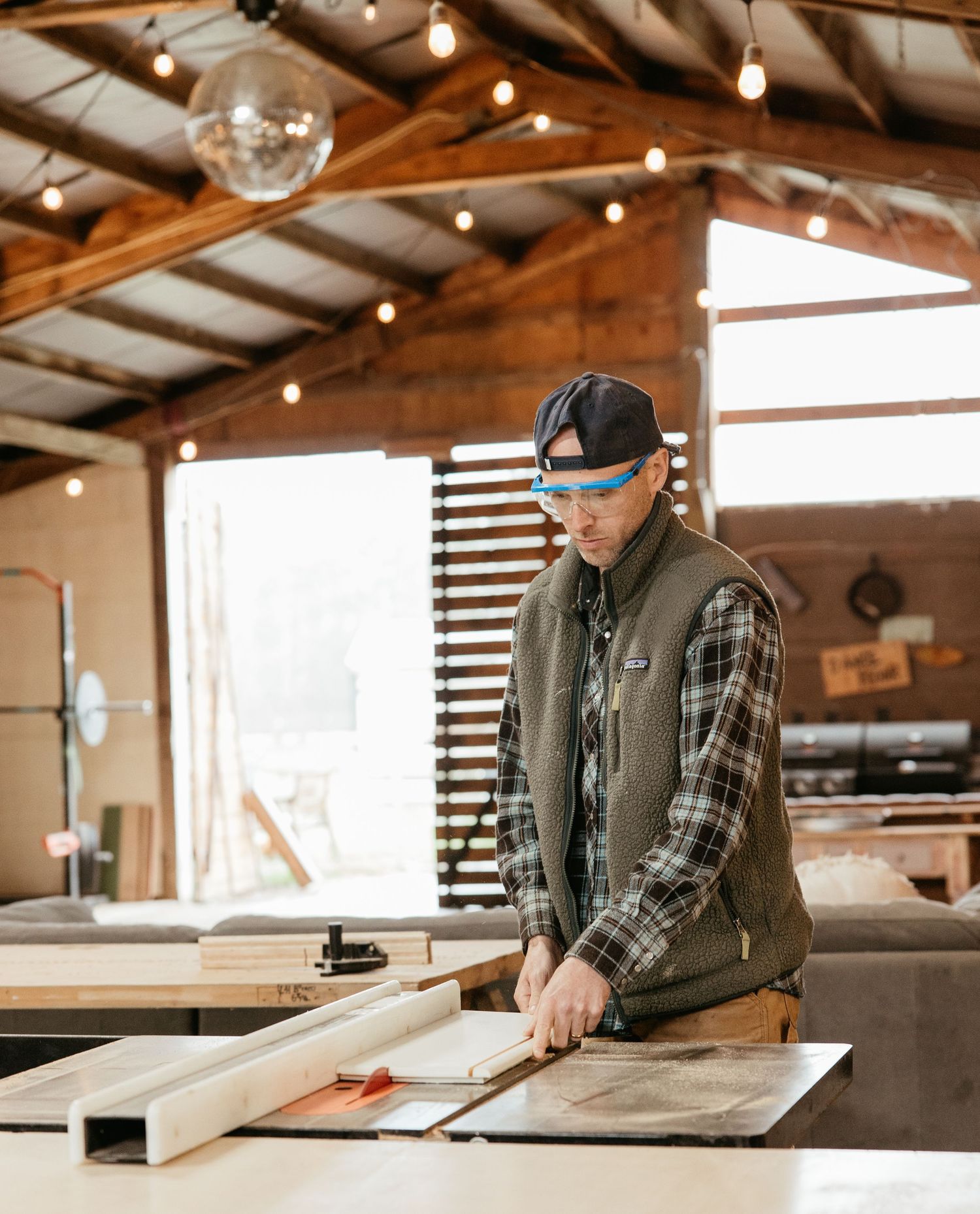 Woodworker in plaid shirt and vest examines plans while working in a sunlit workshop with string lights overhead.