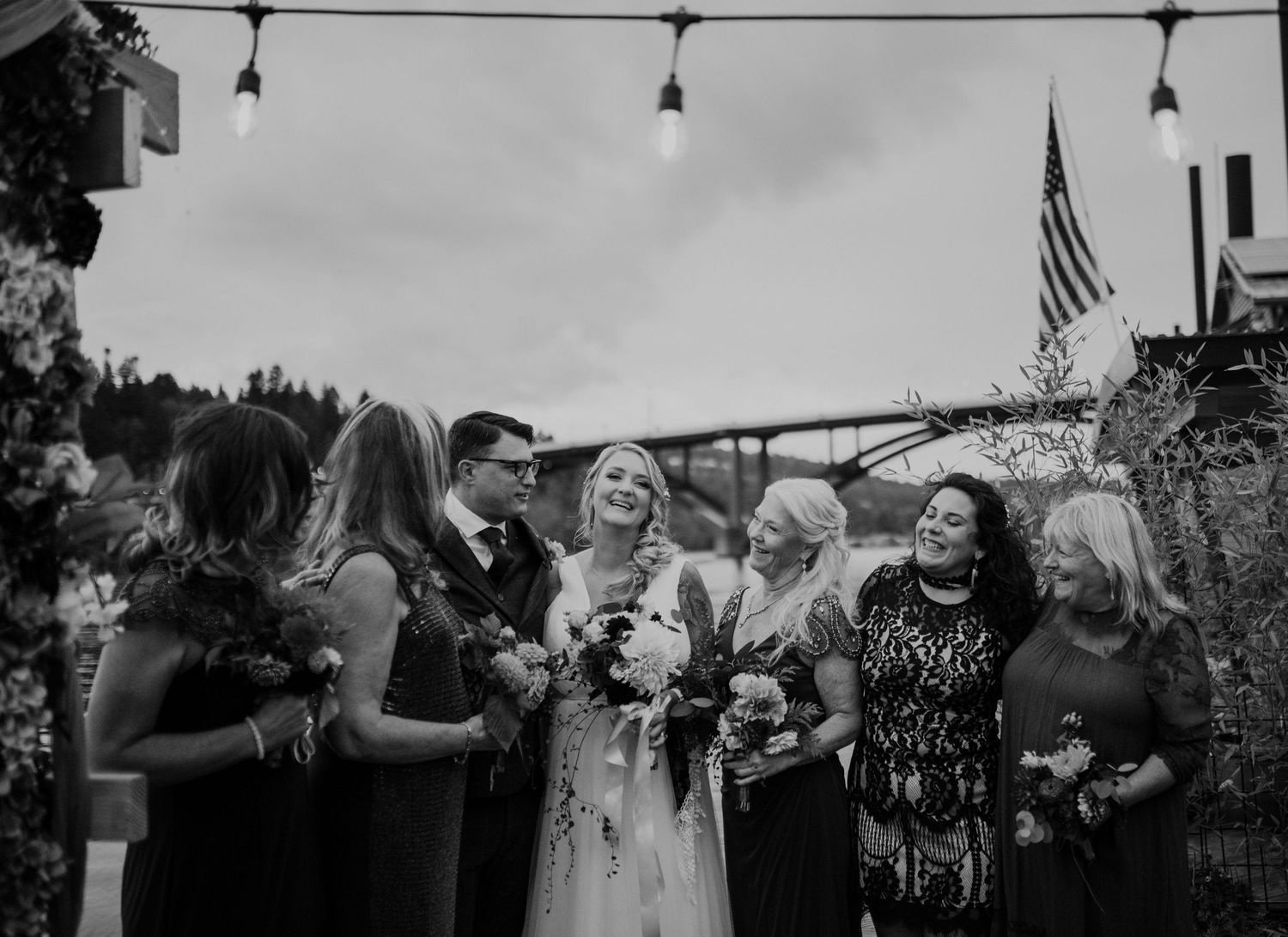 Wedding party celebrates outdoors with string lights and American flag in background.