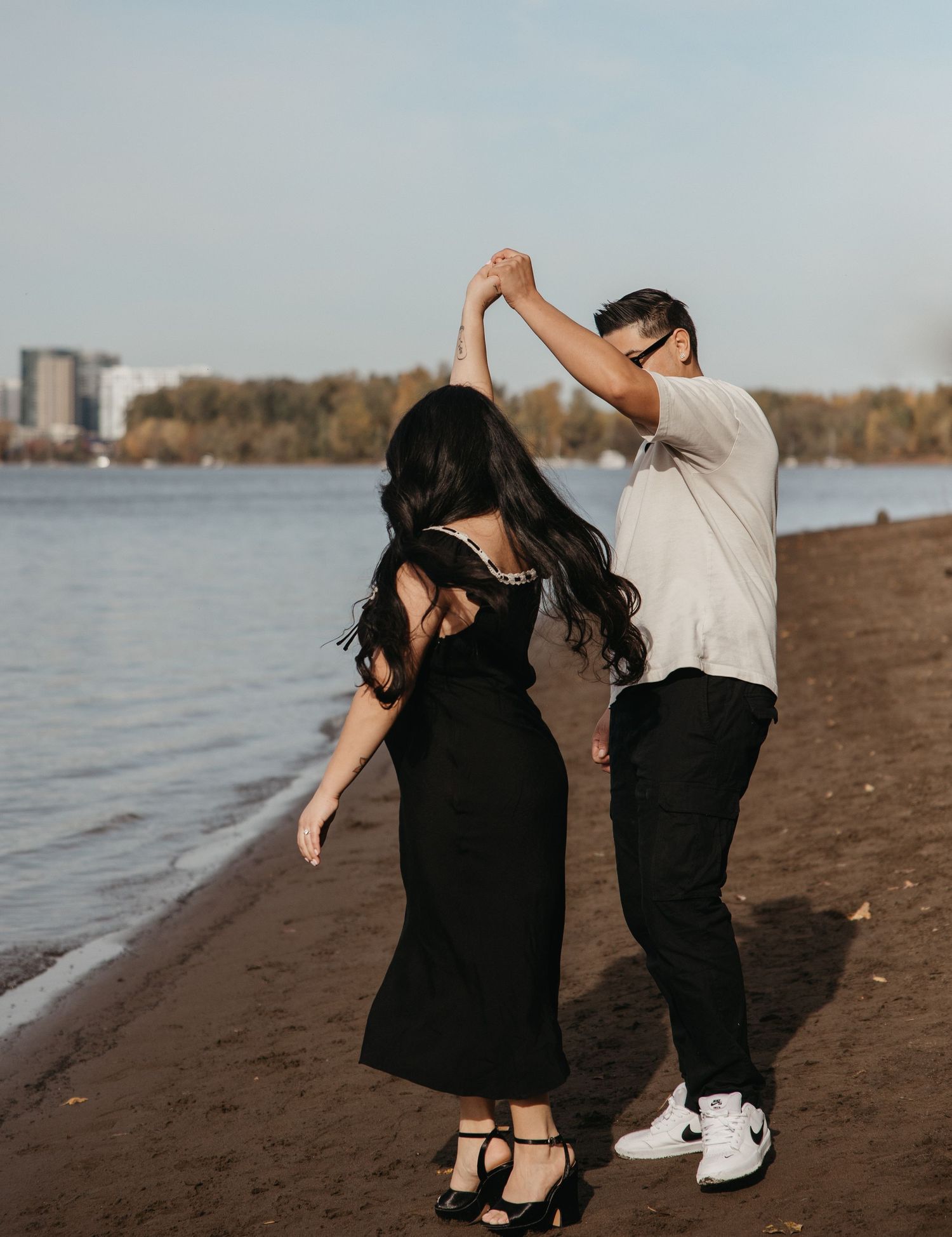 A couple in black and white outfits dance playfully together on a sandy beach at sunset.