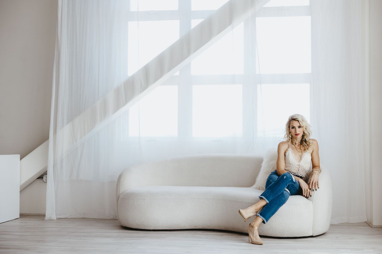 Woman in white dress sits elegantly in bright minimalist studio interior with sunlit windows and flowing sheer curtains.
