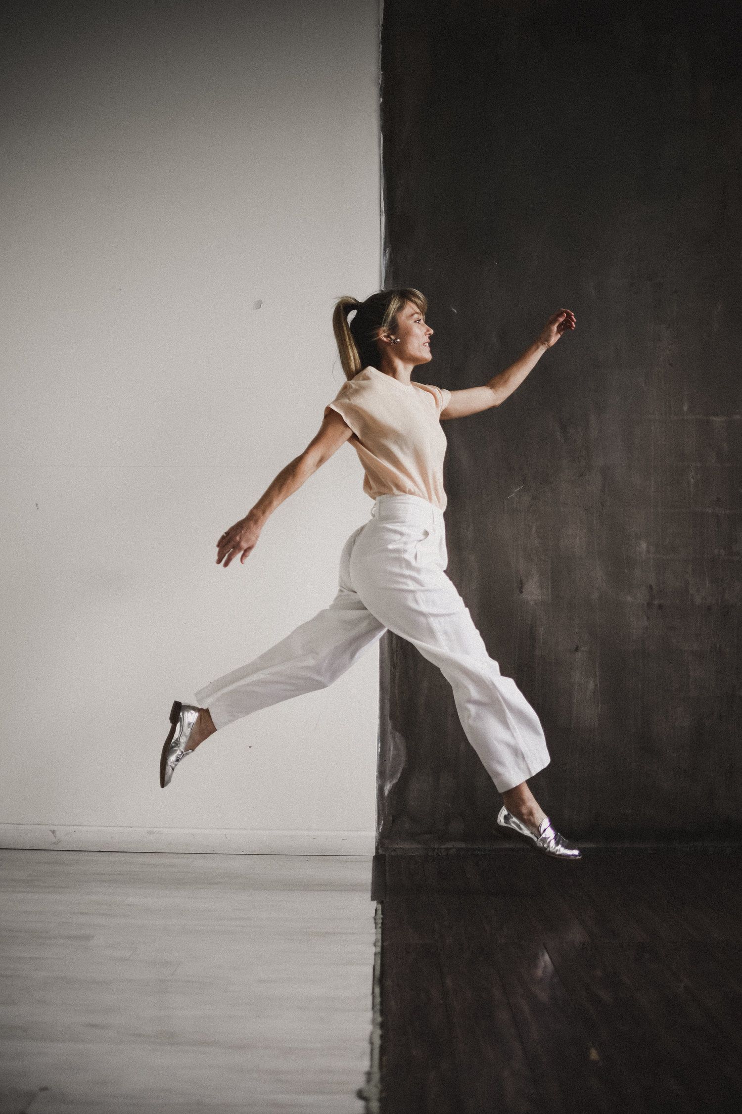 A dancer in white clothing leaps gracefully against a dark backdrop with arms outstretched and one leg extended.