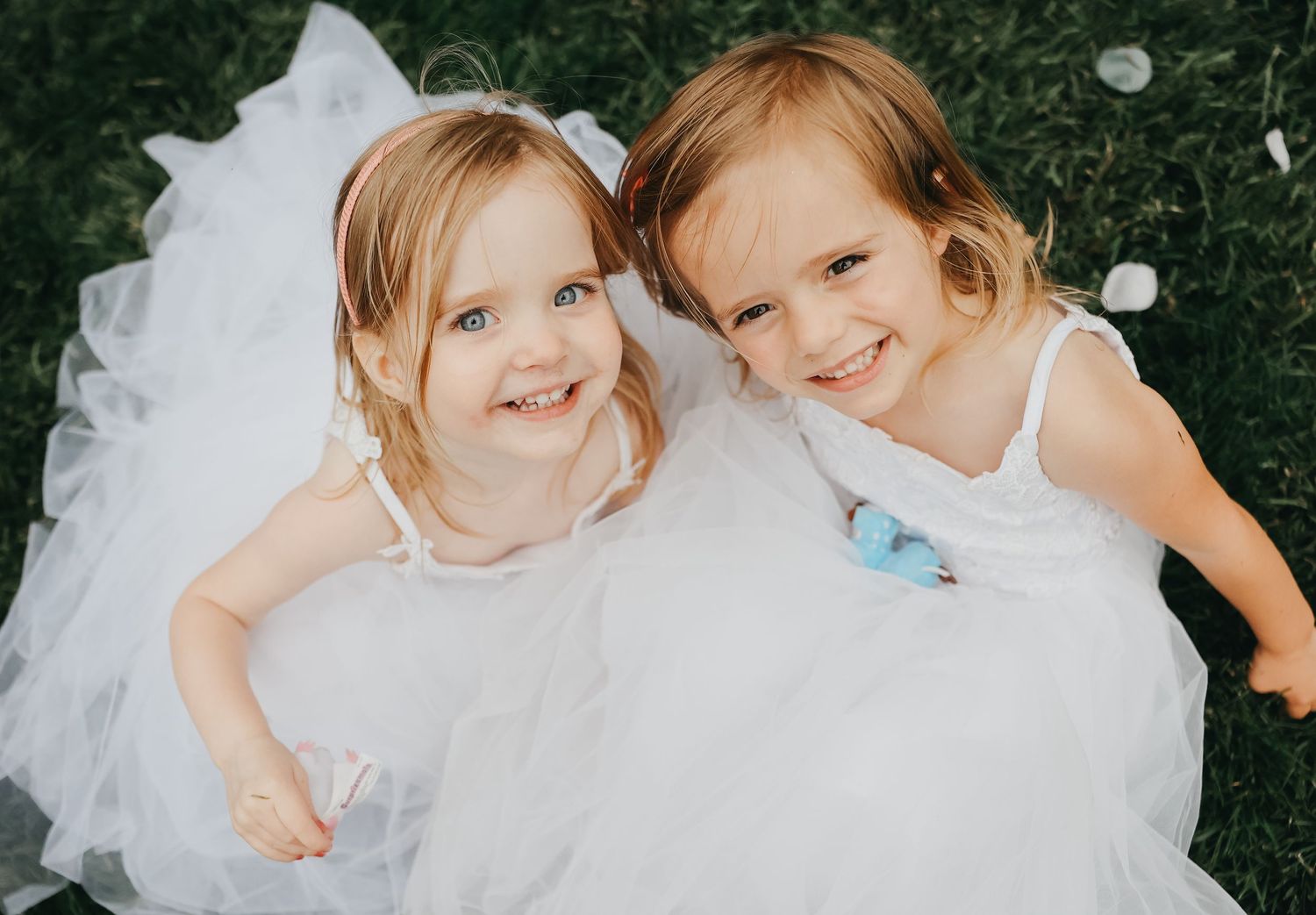 Two young flower girls in white dresses smile joyfully on the grass.