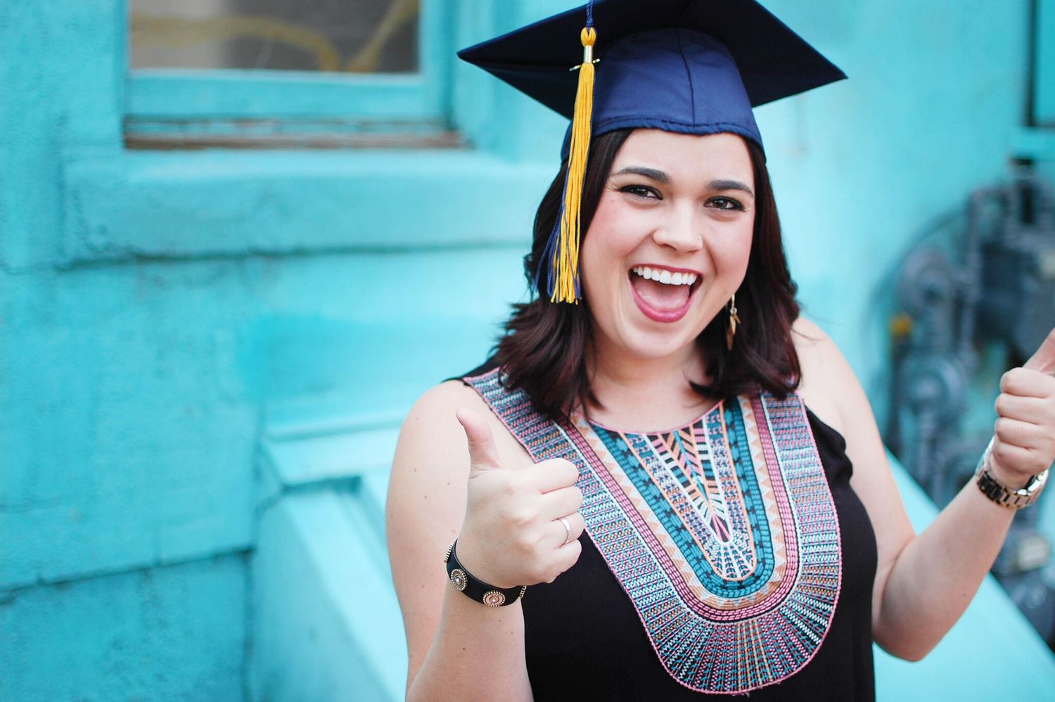 Graduate in cap and gown wearing colorful beaded necklace celebrates with thumbs up against turquoise wall.