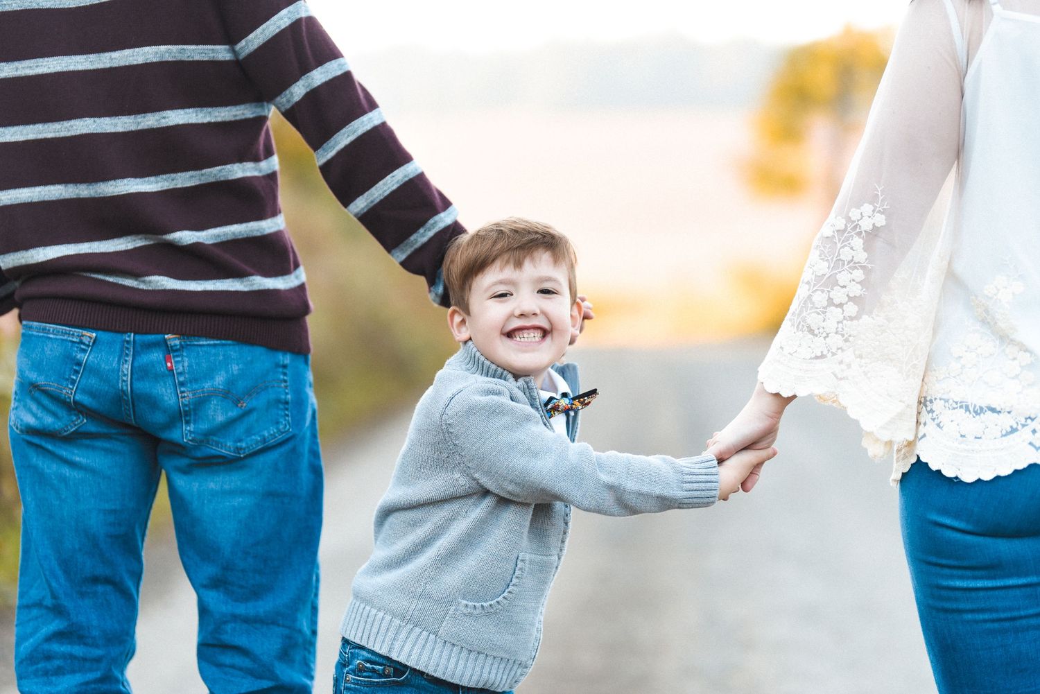 A small child in a gray sweater laughs while holding hands with two adults during a sunset photo session.