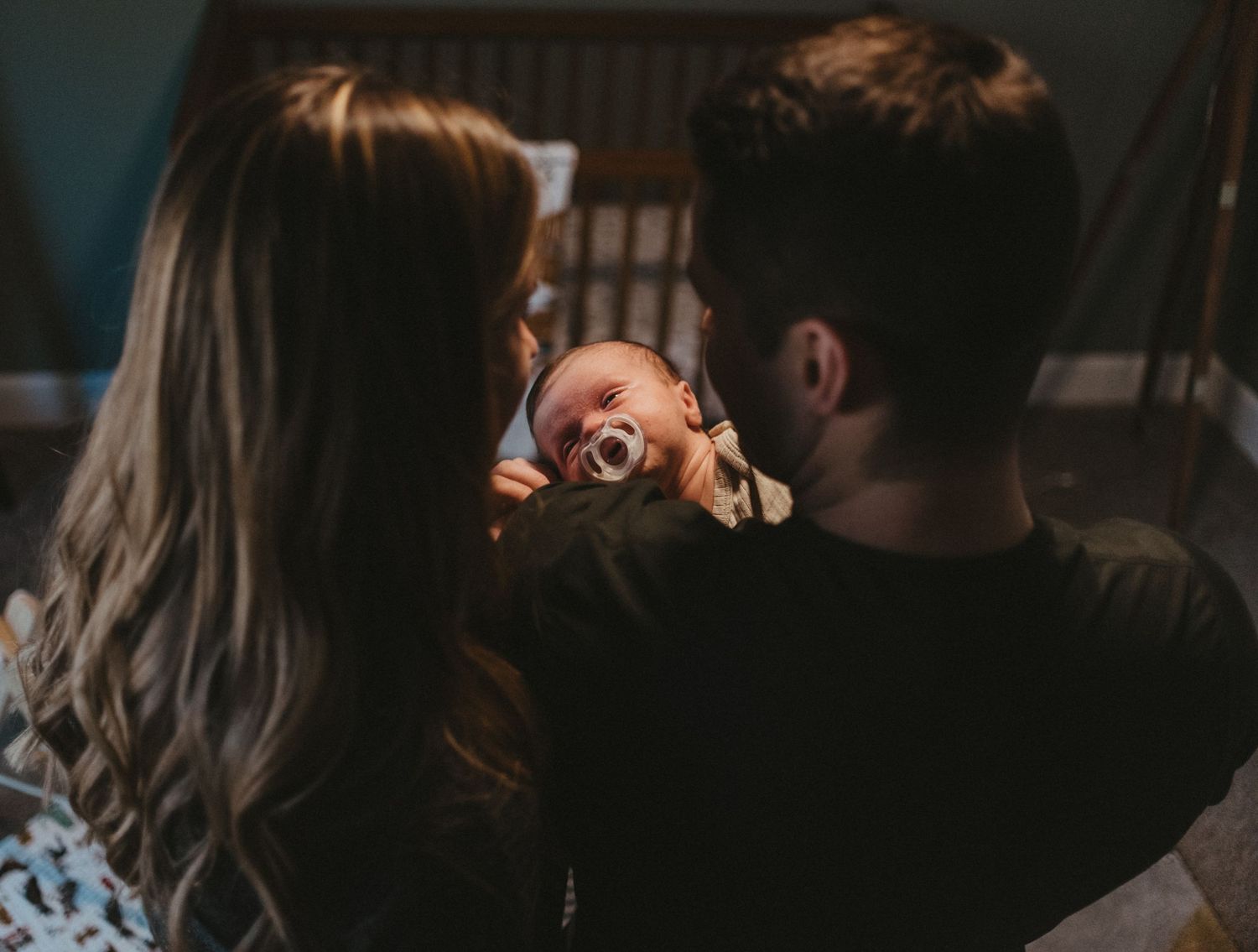 Parents share tender moment with laughing baby in dimly lit room.