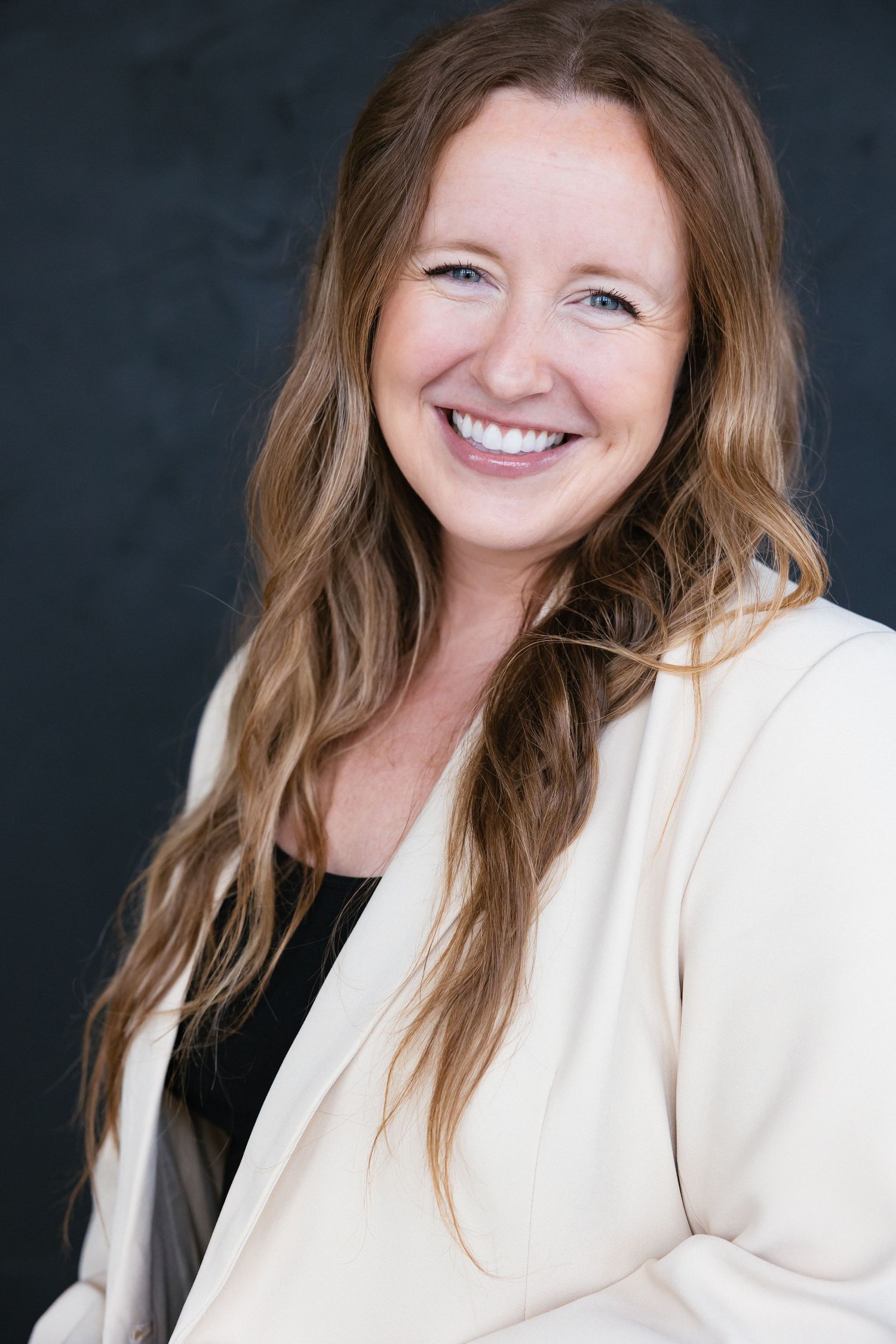 Smiling woman with long wavy hair wearing a cream blazer poses for a professional headshot against a dark background.