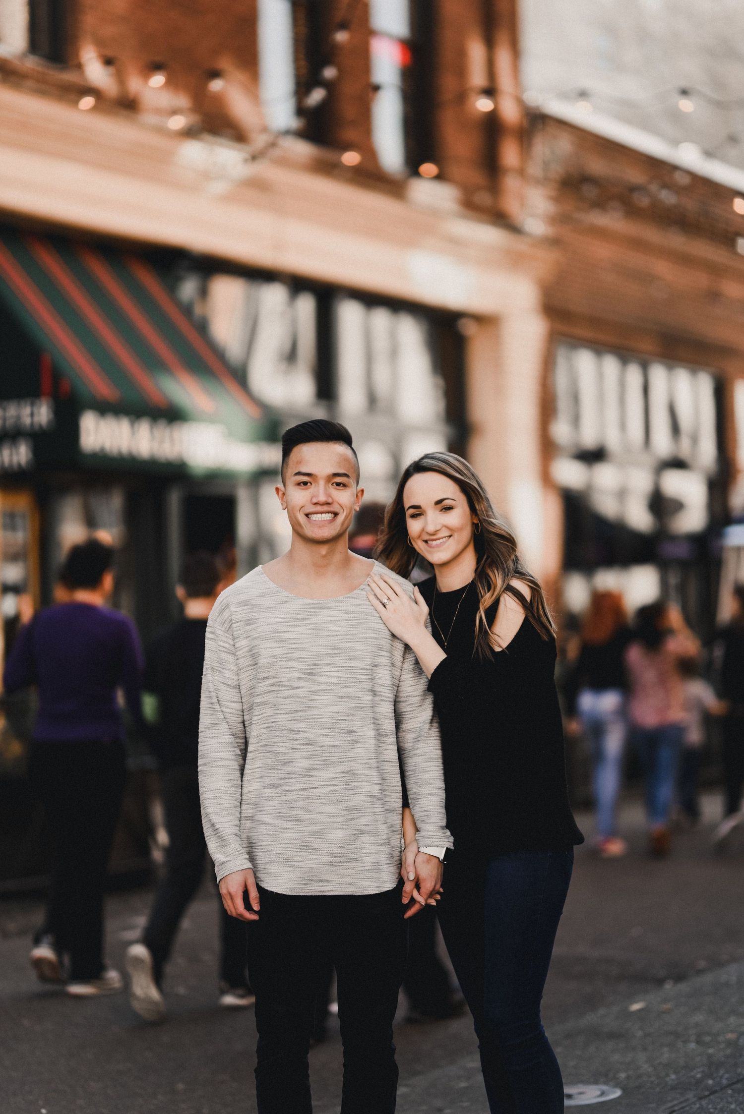 A couple smiles together while standing on a city sidewalk near storefronts during daytime.