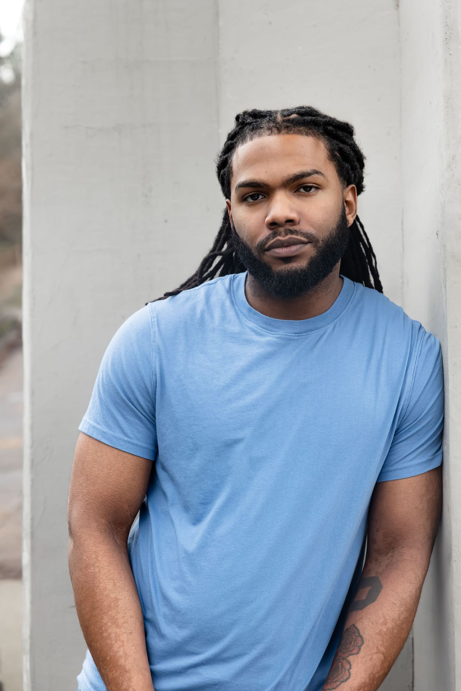 Young Black man with dreadlocks and beard wearing a blue t-shirt, leaning against a concrete wall outdoors.