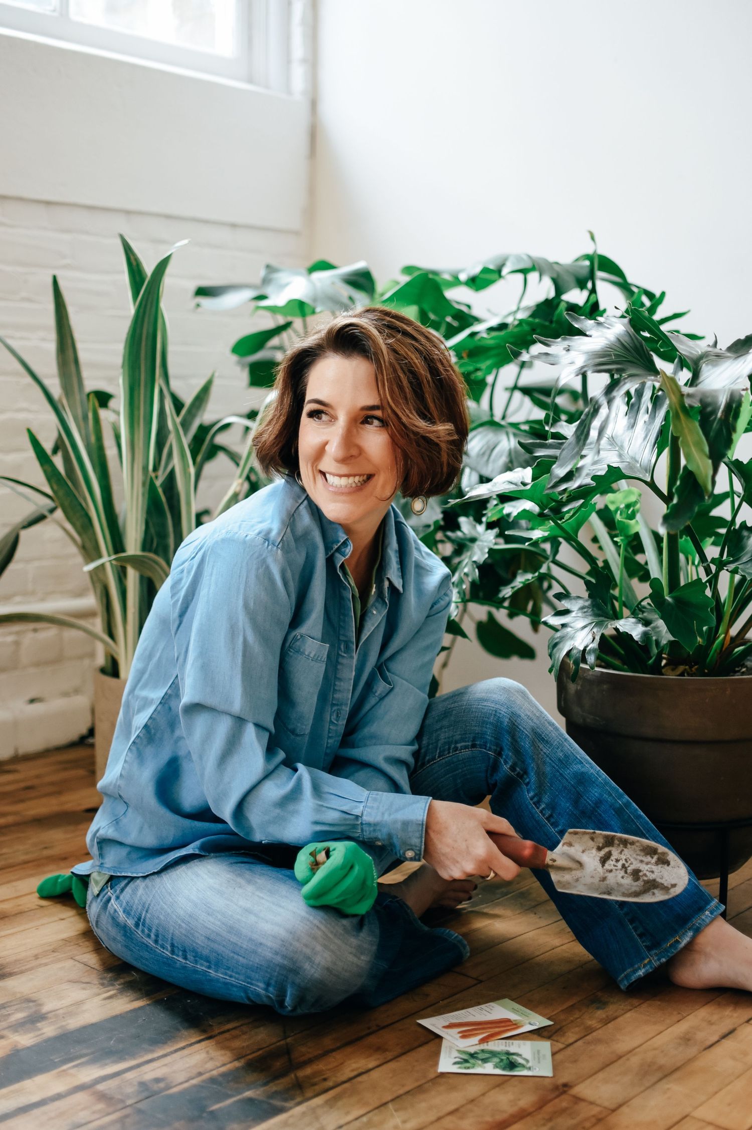 A person in denim relaxes on a hardwood floor surrounded by potted plants in a bright, modern interior space.