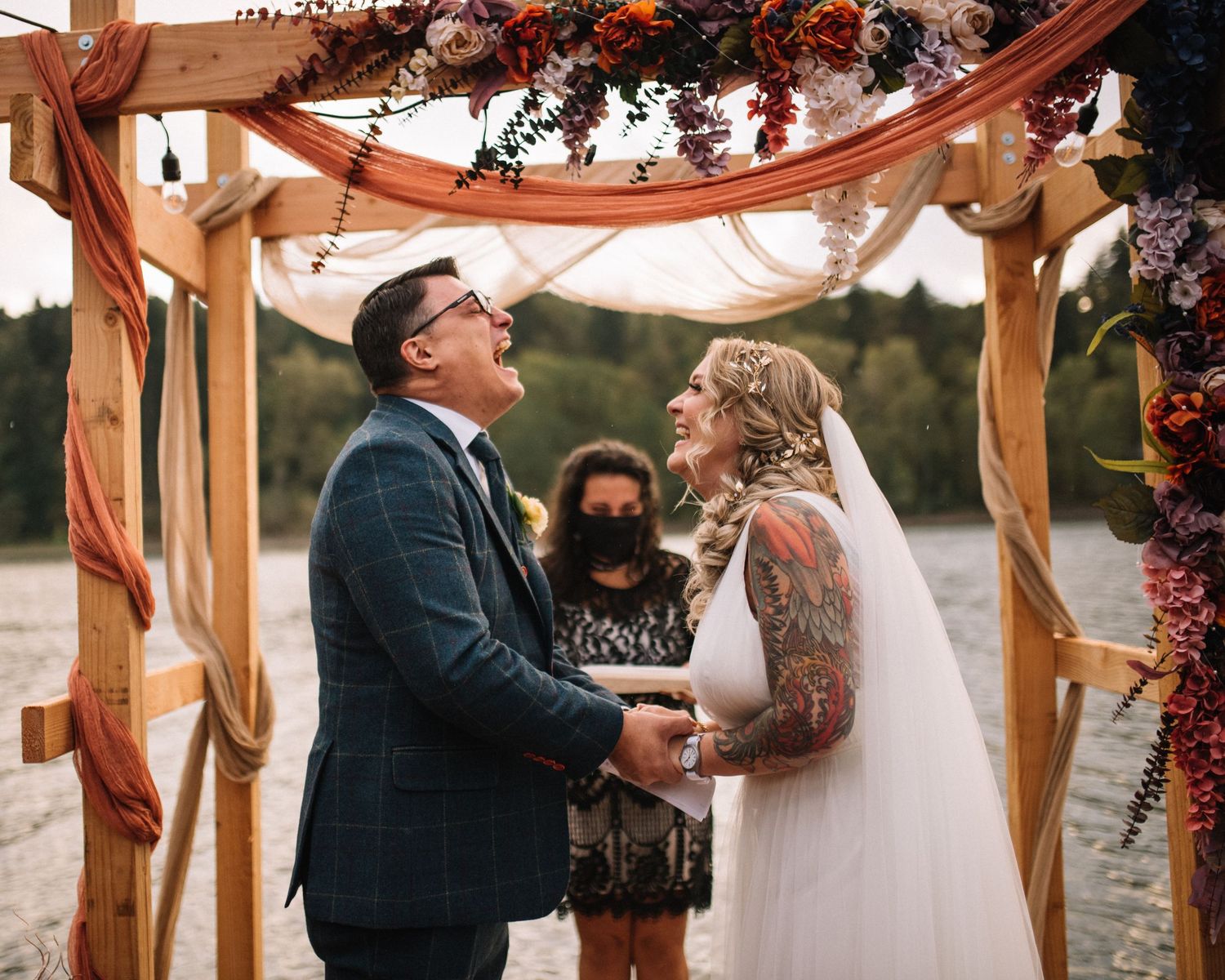 A joyful couple exchanges vows under a wooden arch decorated with orange and purple flowers.