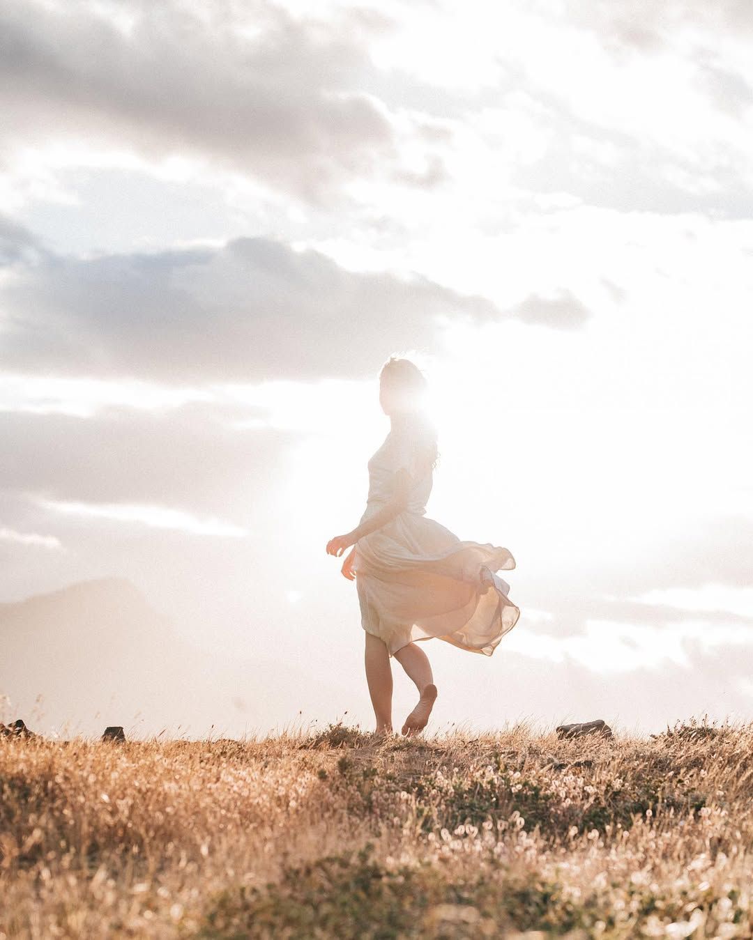 A silhouetted figure stands in a flowing dress against a sunset sky on a hilltop.