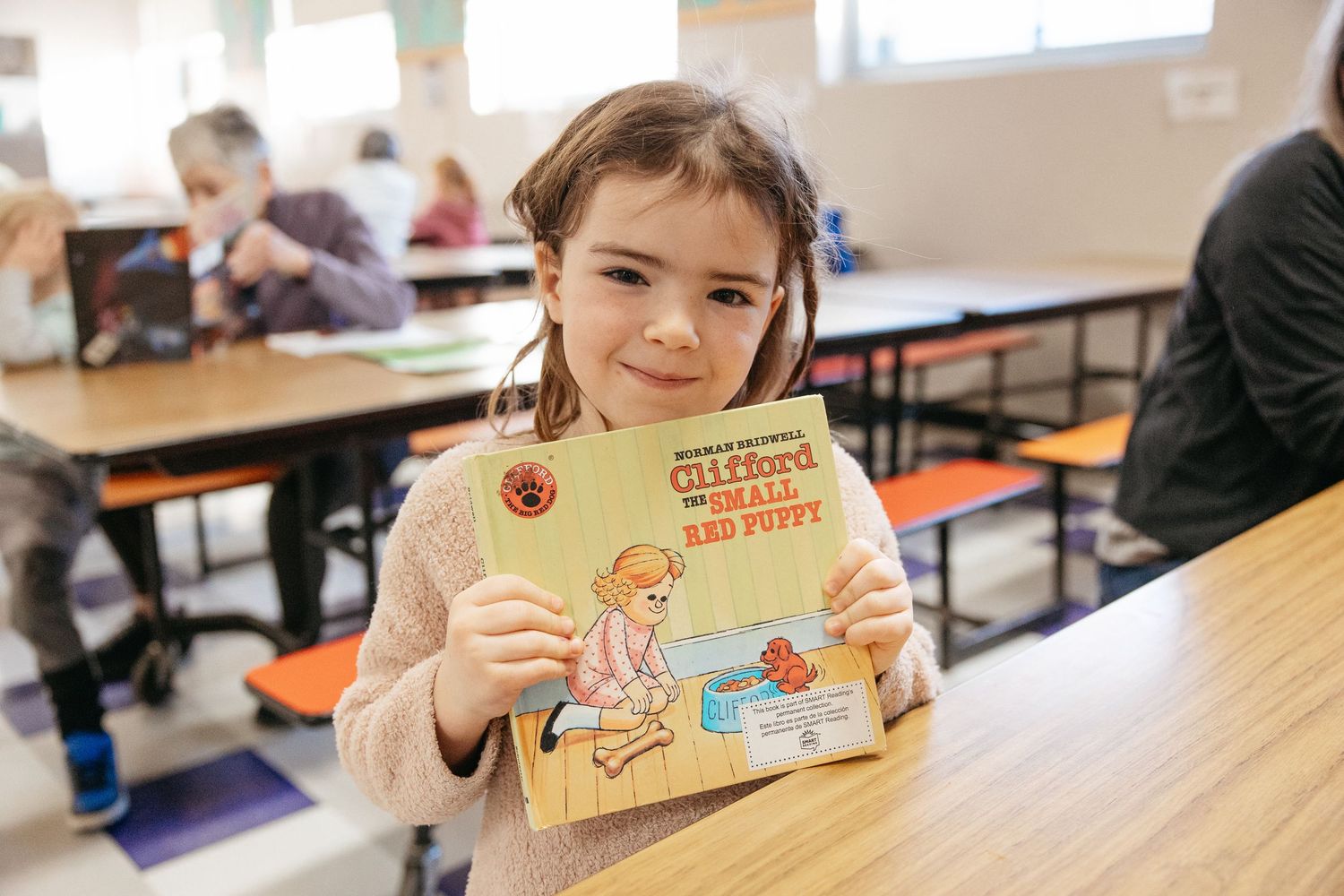A student in a classroom holds up a children's book while sitting at a desk.