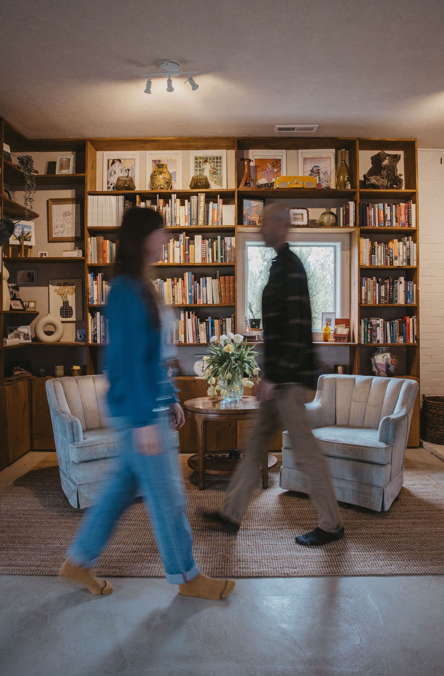 Two figures in motion create a blurred effect while dancing through a cozy home library with bookshelves and armchairs.