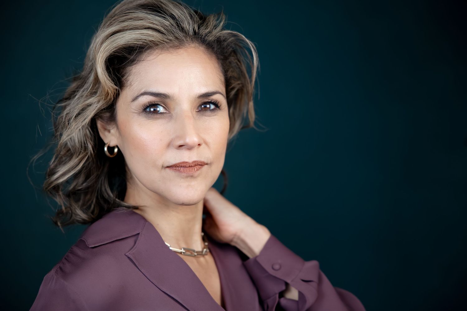 Professional headshot of a confident woman with wavy gray-streaked hair wearing a mauve blazer against teal background.