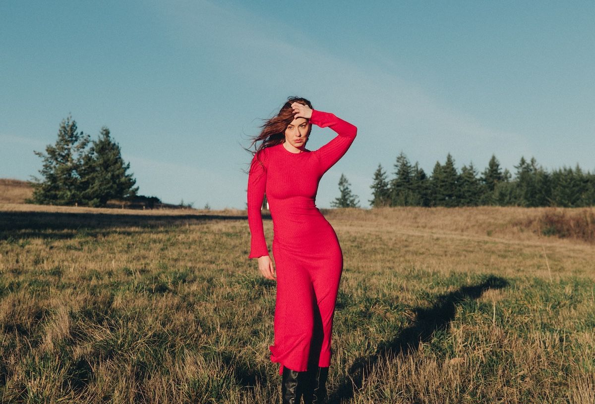 A figure in a vibrant red long-sleeved dress stands in a golden field at sunset with evergreen trees in the background.