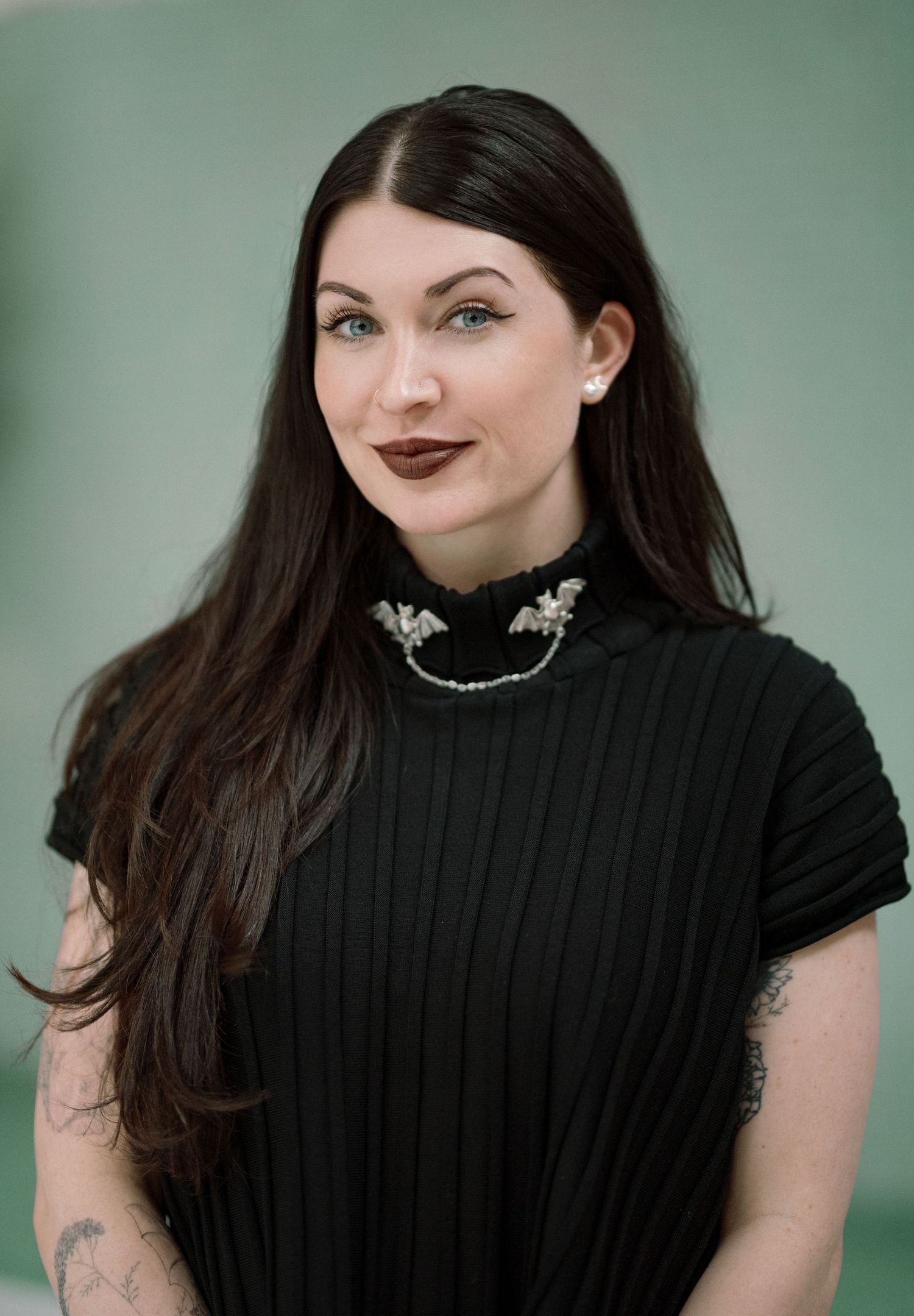 Woman with long dark hair wearing a black ribbed top with bat brooch and visible tattoos, posing for a professional portrait.