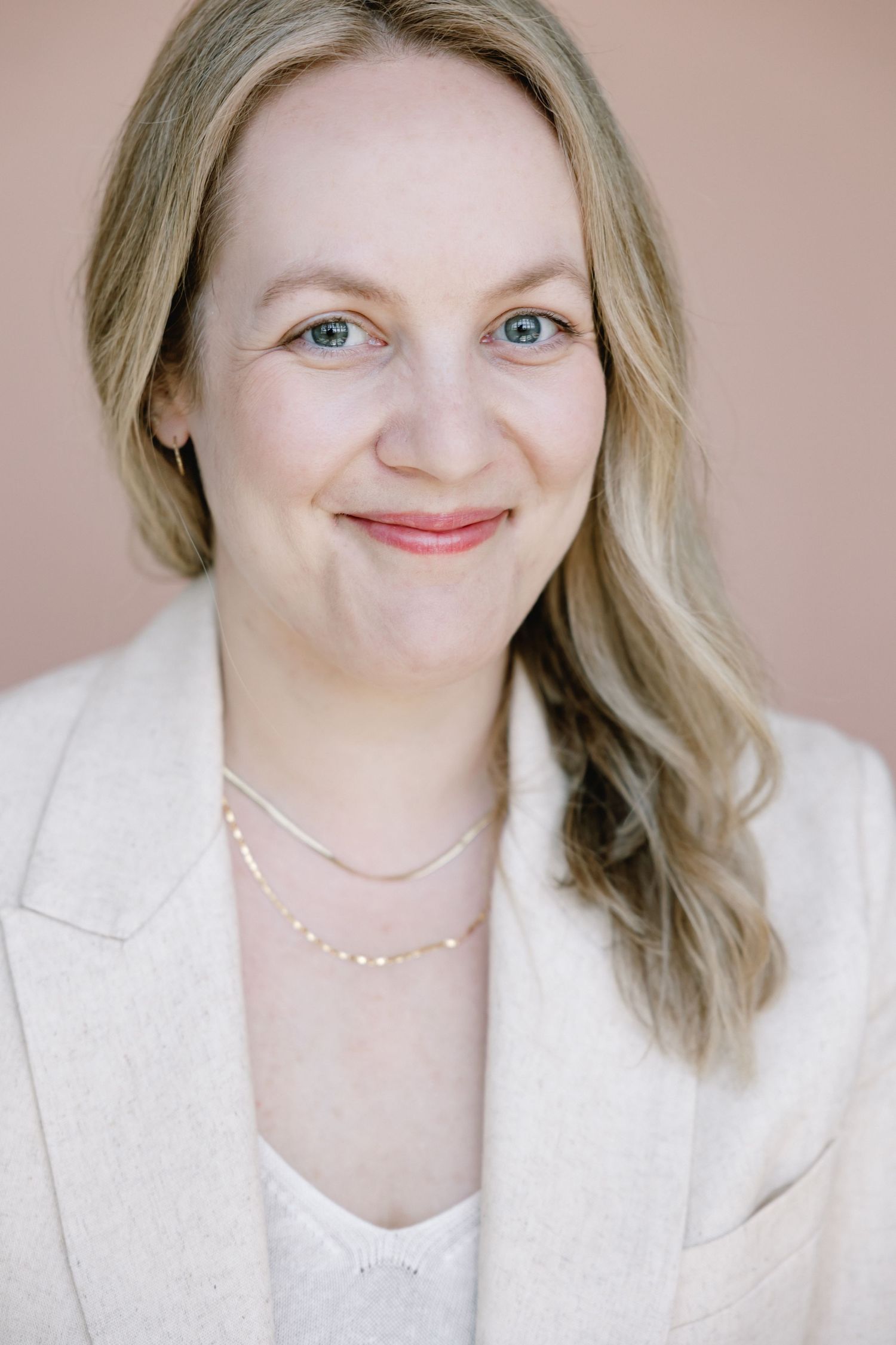 Smiling blonde woman with blue eyes wearing a cream blazer and delicate pearl necklace against a soft pink background.