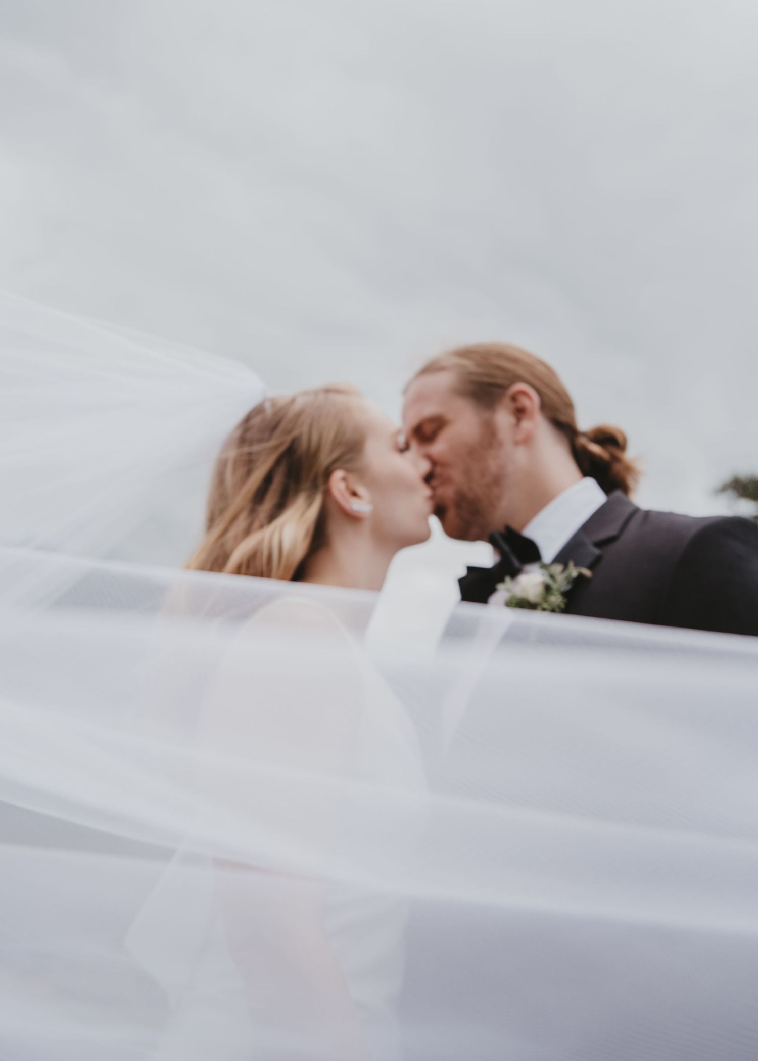 A romantic moment under a flowing white veil against an overcast sky.