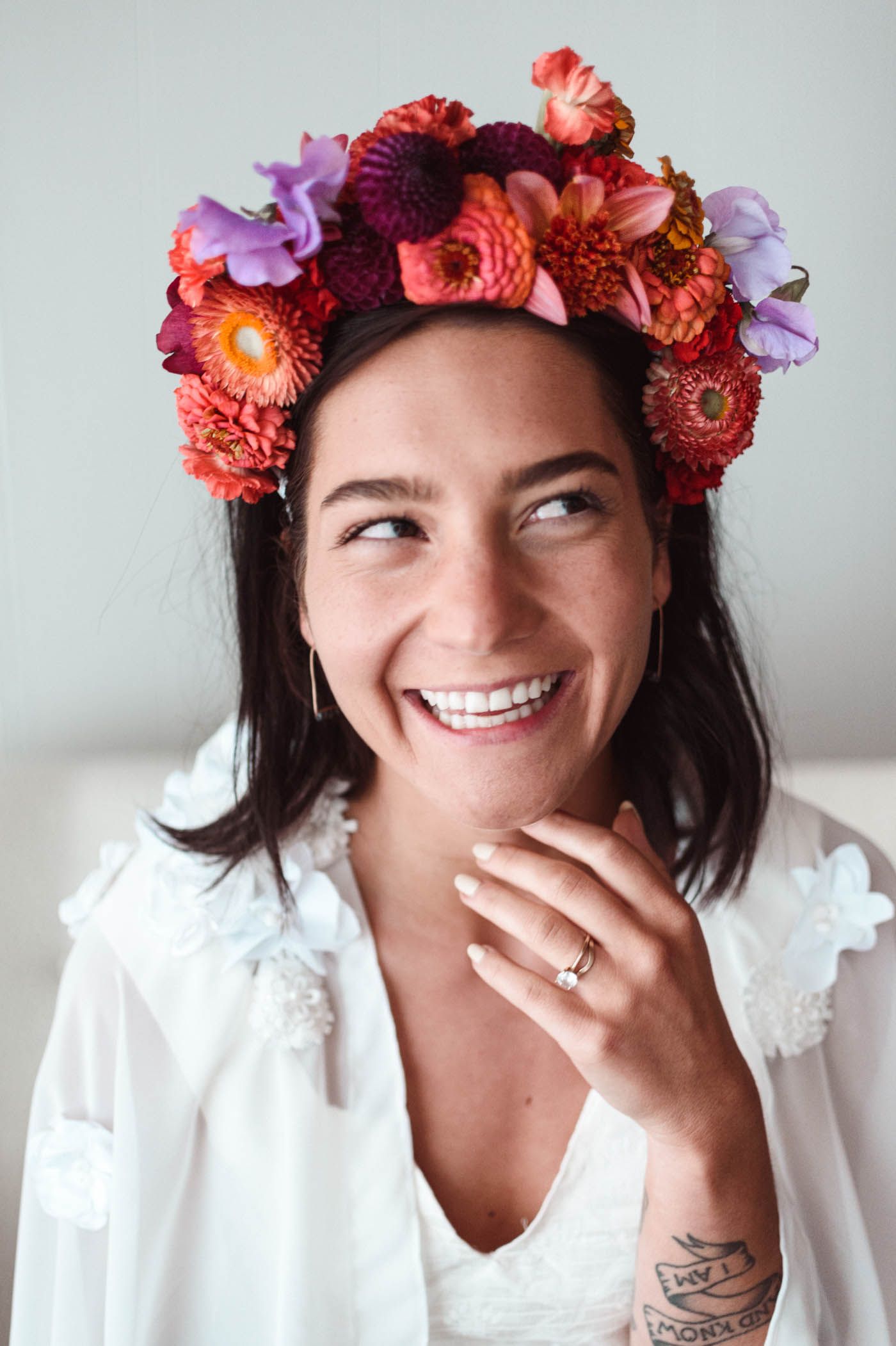 A woman wearing a vibrant floral crown of red and purple blooms smiles while wearing a white top.