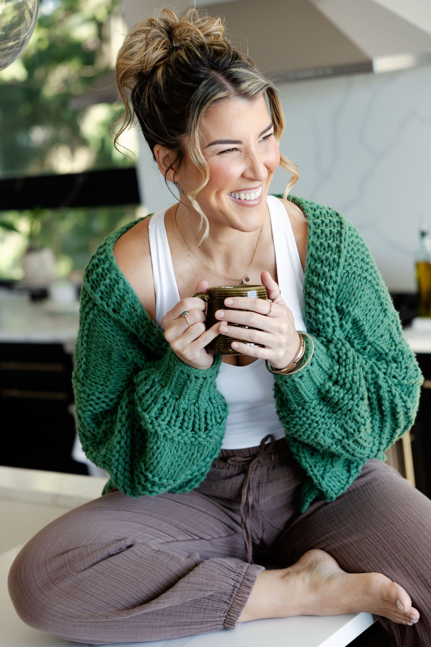 Smiling woman in green knit cardigan sitting cross-legged holding a green mug, laughing joyfully indoors.