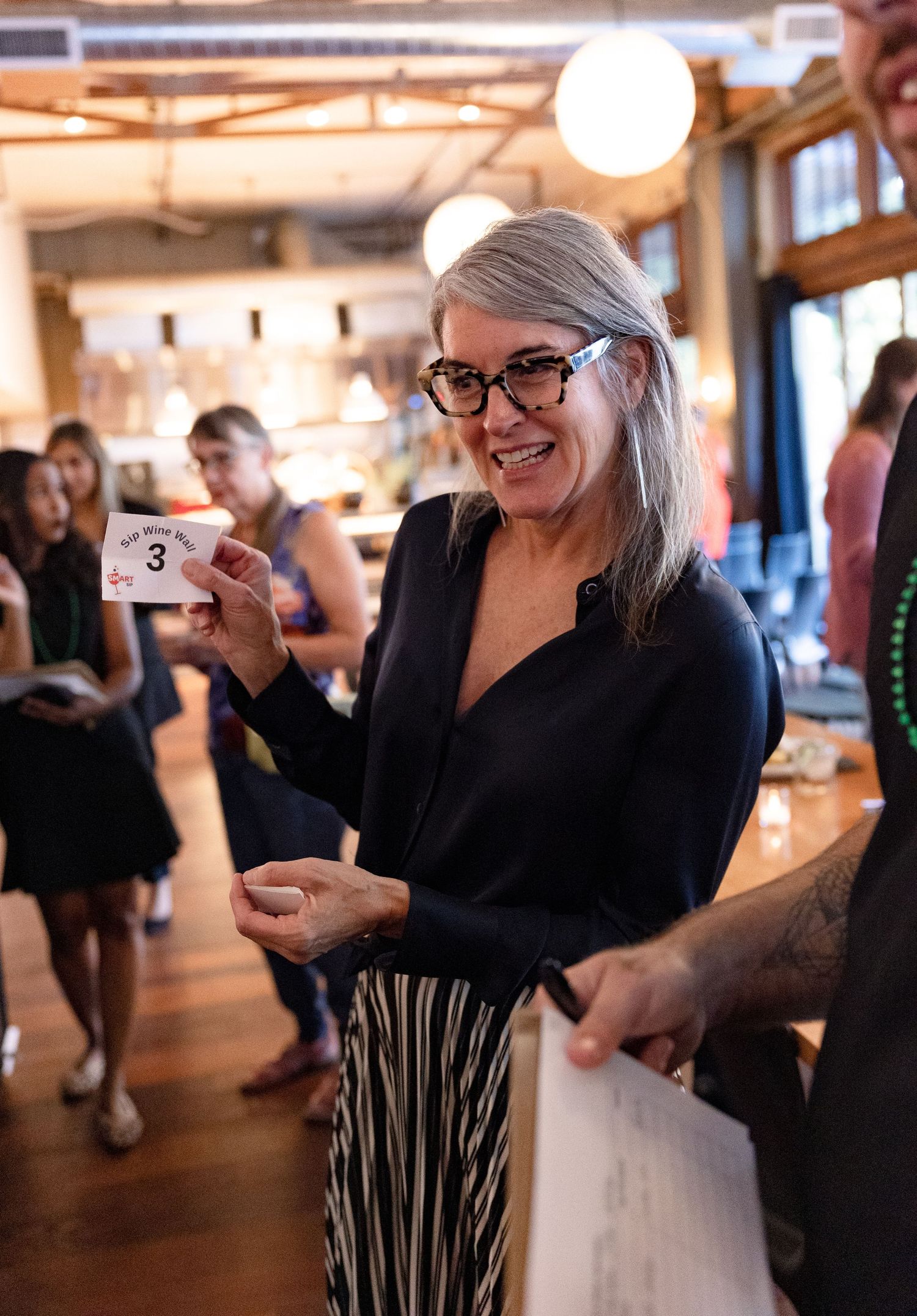 A woman in a black top and striped skirt smiles while holding a numbered card at an indoor social gathering.