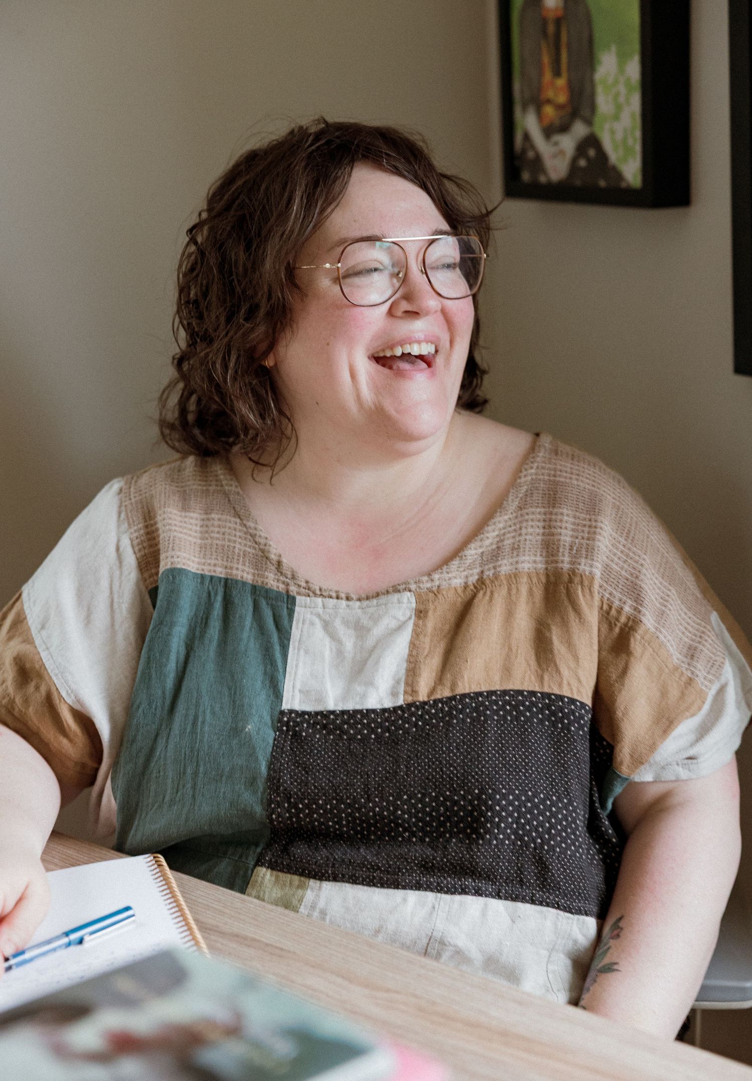 A smiling woman with curly hair and glasses wearing a colorful patchwork top sits at a desk laughing.